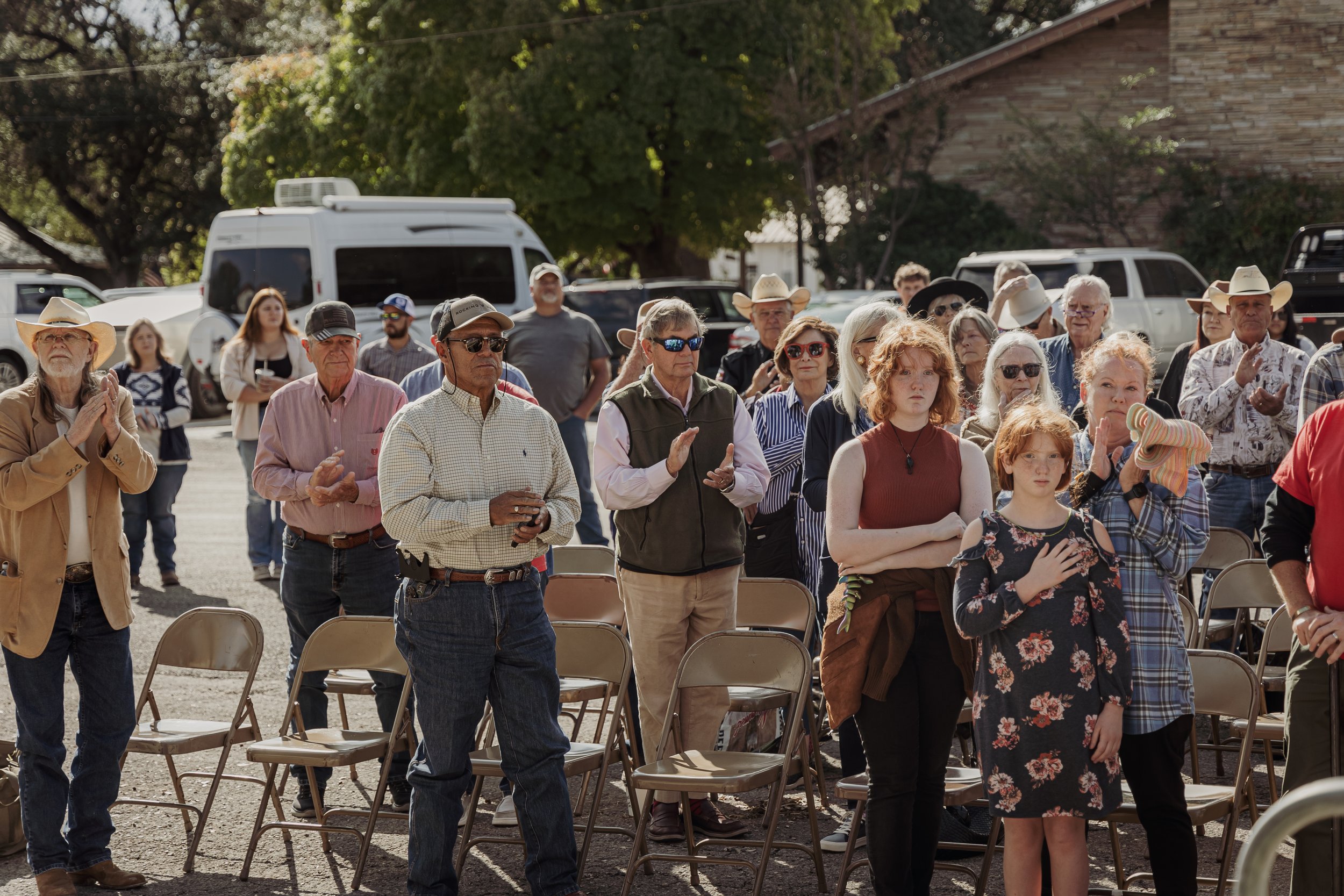 A group of people standing outdoors during a daytime event, some with their hands over their hearts and others clapping, with chairs and vehicles in the background.