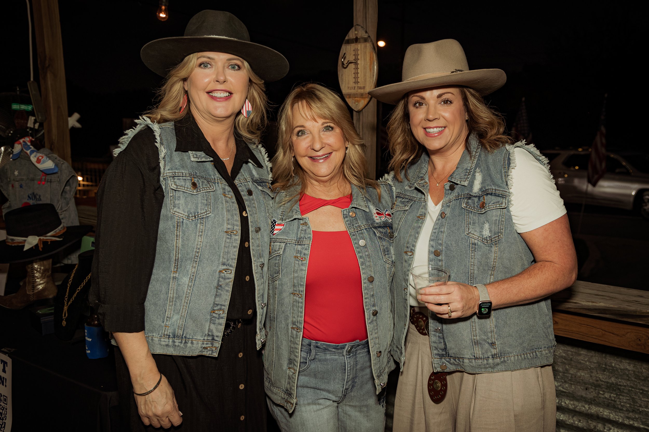 Three women wearing denim vests and hats, smiling, at a nighttime outdoor event with patriotic decorations.