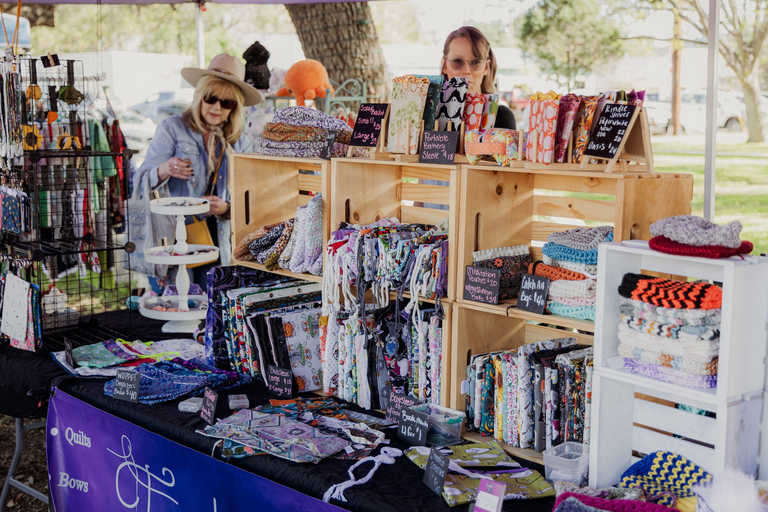 Diverse handmade crafts, such as knit hats, fabric pouches, bags, and quilts, displayed on tables and shelves at an outdoor market stall with two women browsing.