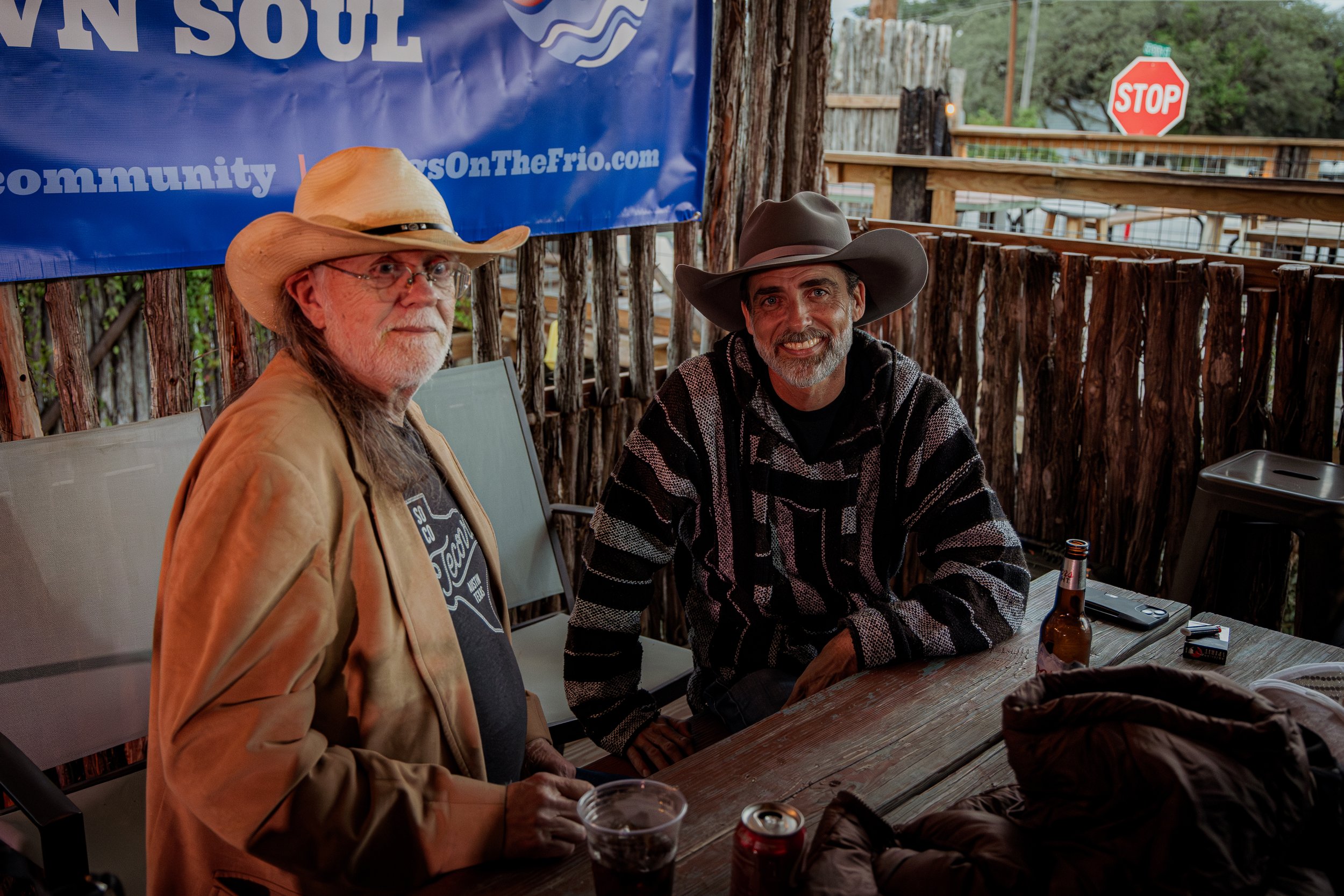 Two men wearing cowboy hats sitting at a wooden table outdoors, smiling at the camera, with drinks and a banner in the background.