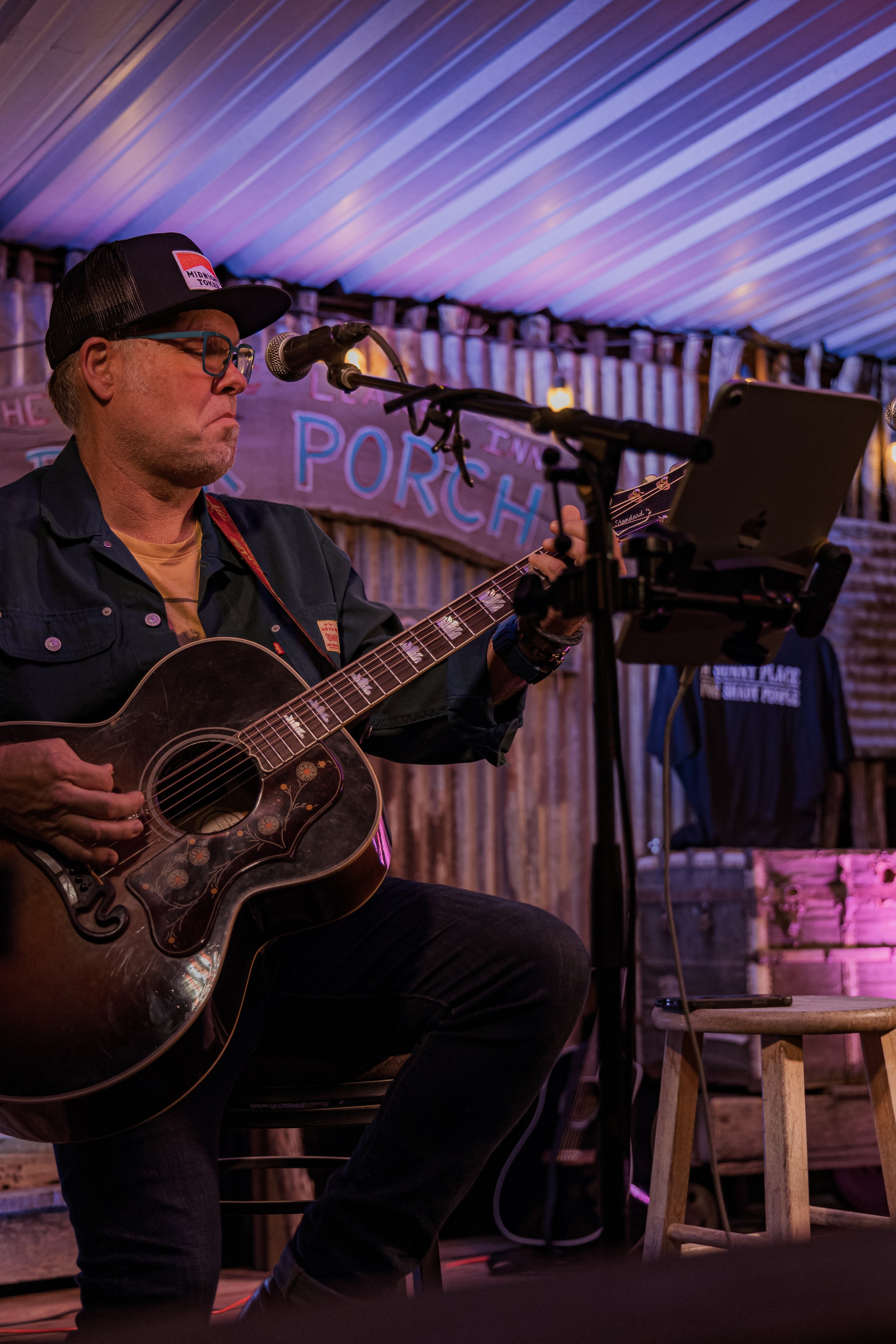 A man wearing glasses, a baseball cap, and a dark shirt playing an acoustic guitar while sitting on a stool on a stage with wood paneling background. There is a microphone stand and a tablet on a stand in front of him. The stage has a sign that says