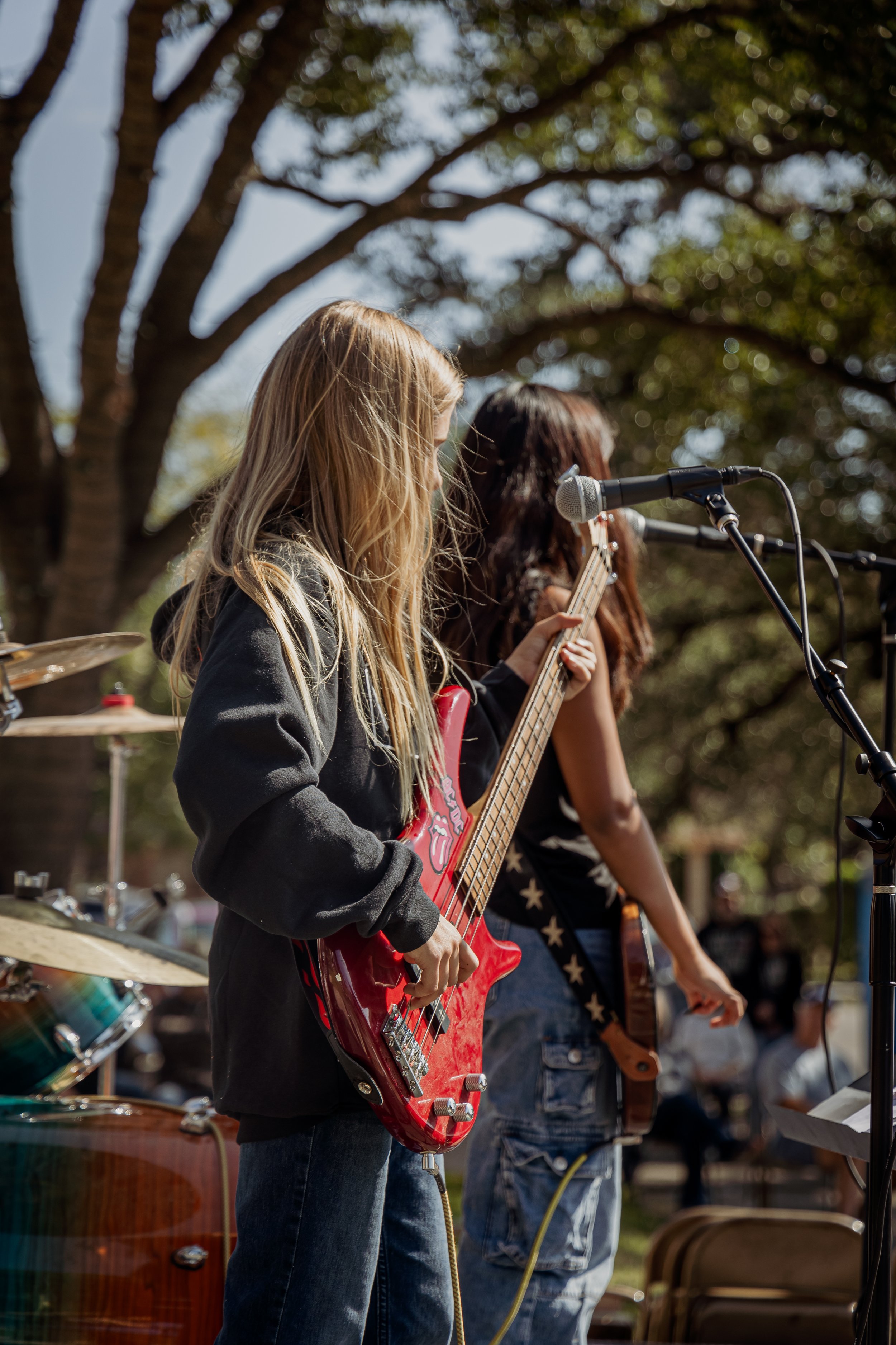 Two young women perform music outdoors, one playing a red bass guitar and the other singing or playing a guitar, with microphones set up, under trees.
