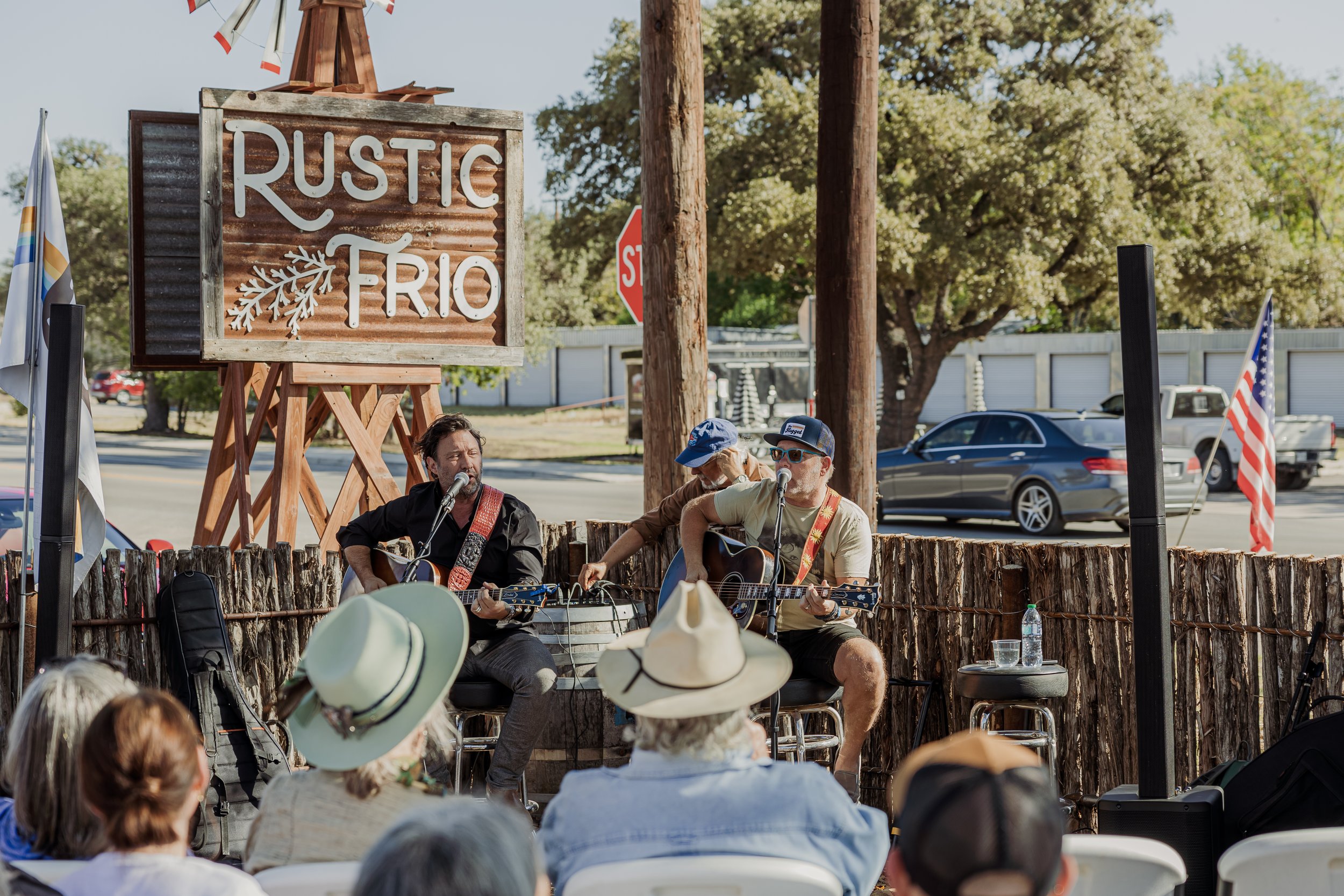 Two musicians perform on an outdoor stage with the sign 'Rustic Trío' in the background, a small audience seated in front, and a backdrop of trees, parked cars, and a clear sky.