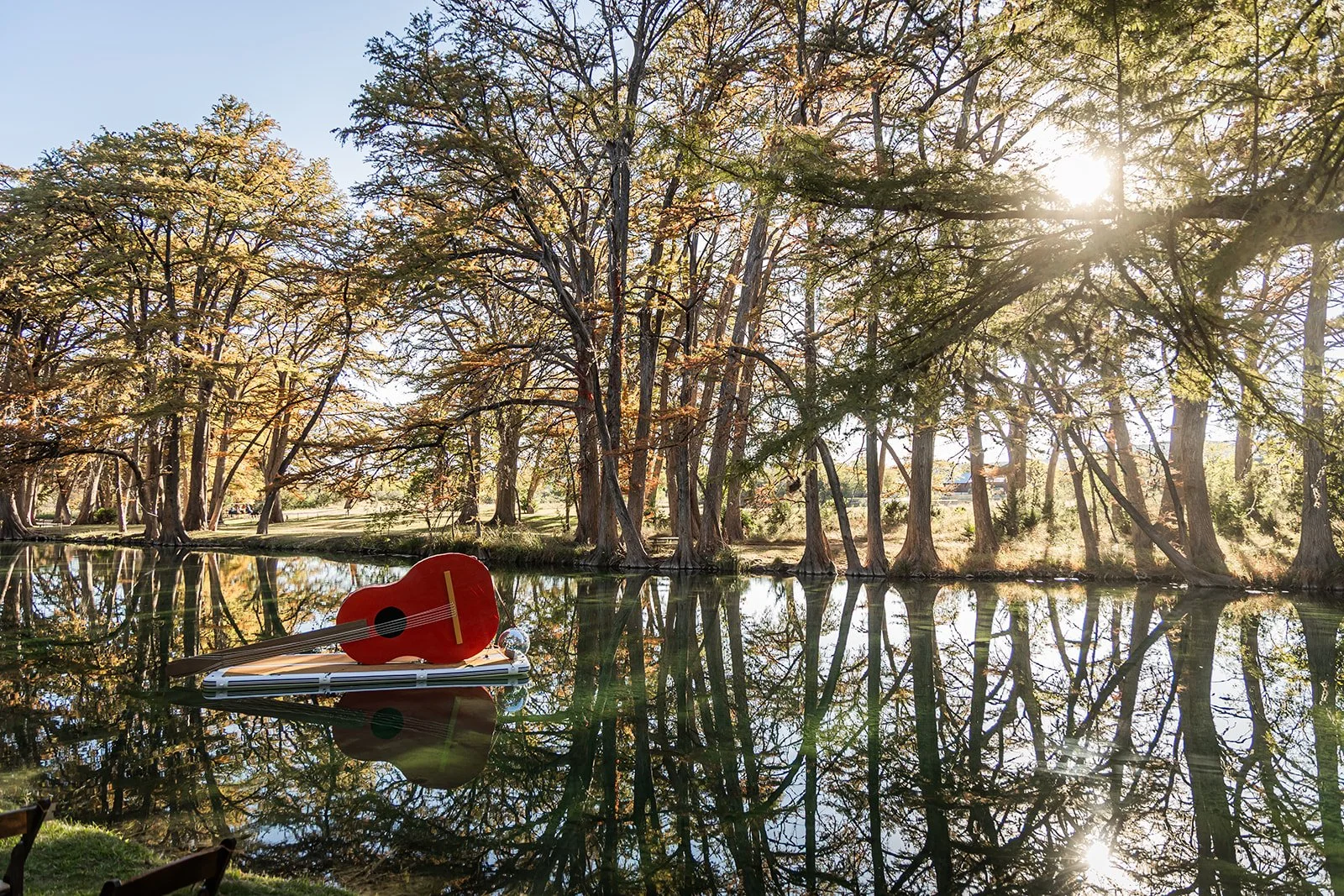 A red ukulele resting on a small boat floating on a calm river, surrounded by tall trees with autumn foliage, reflected in the water, with sunlight filtering through the branches.