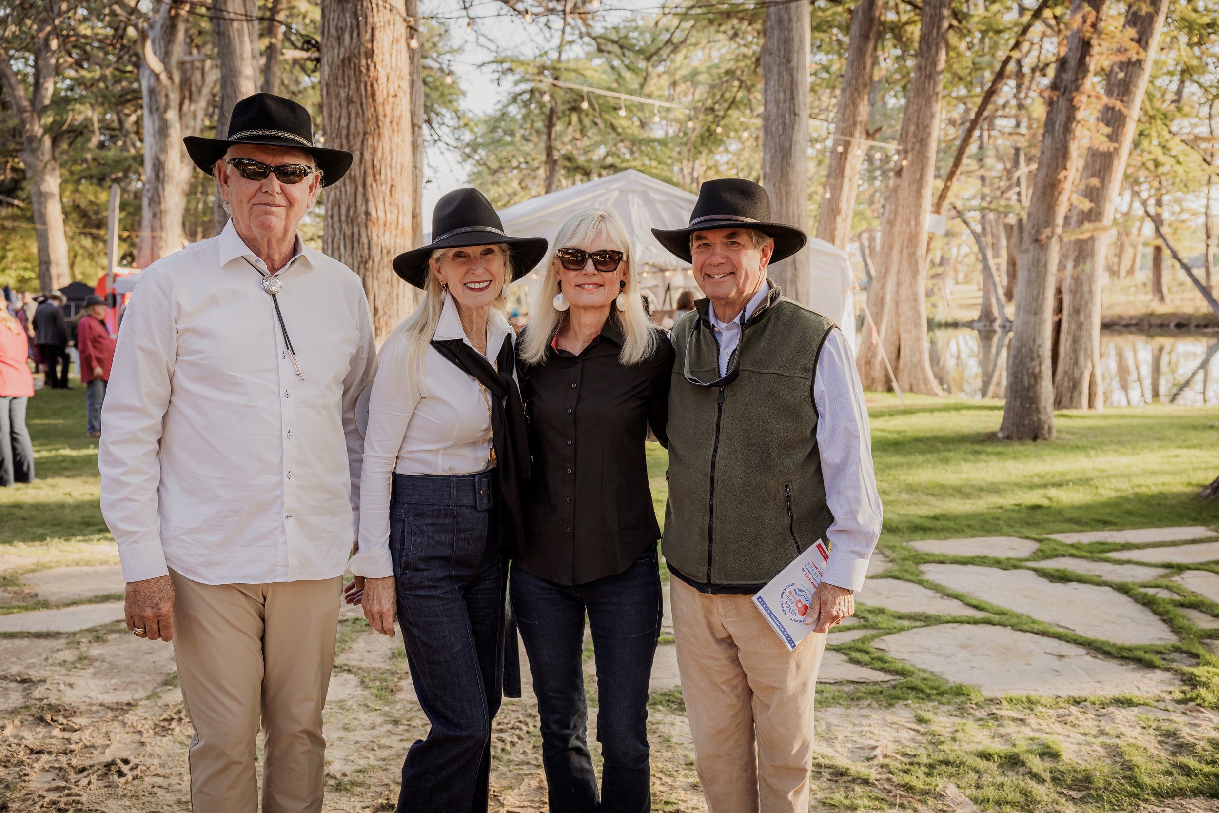 Four older adults standing outdoors in a park with tall trees, smiling at the camera. They are dressed in casual, country-style clothing, with hats and sunglasses, and are gathered for a social event.
