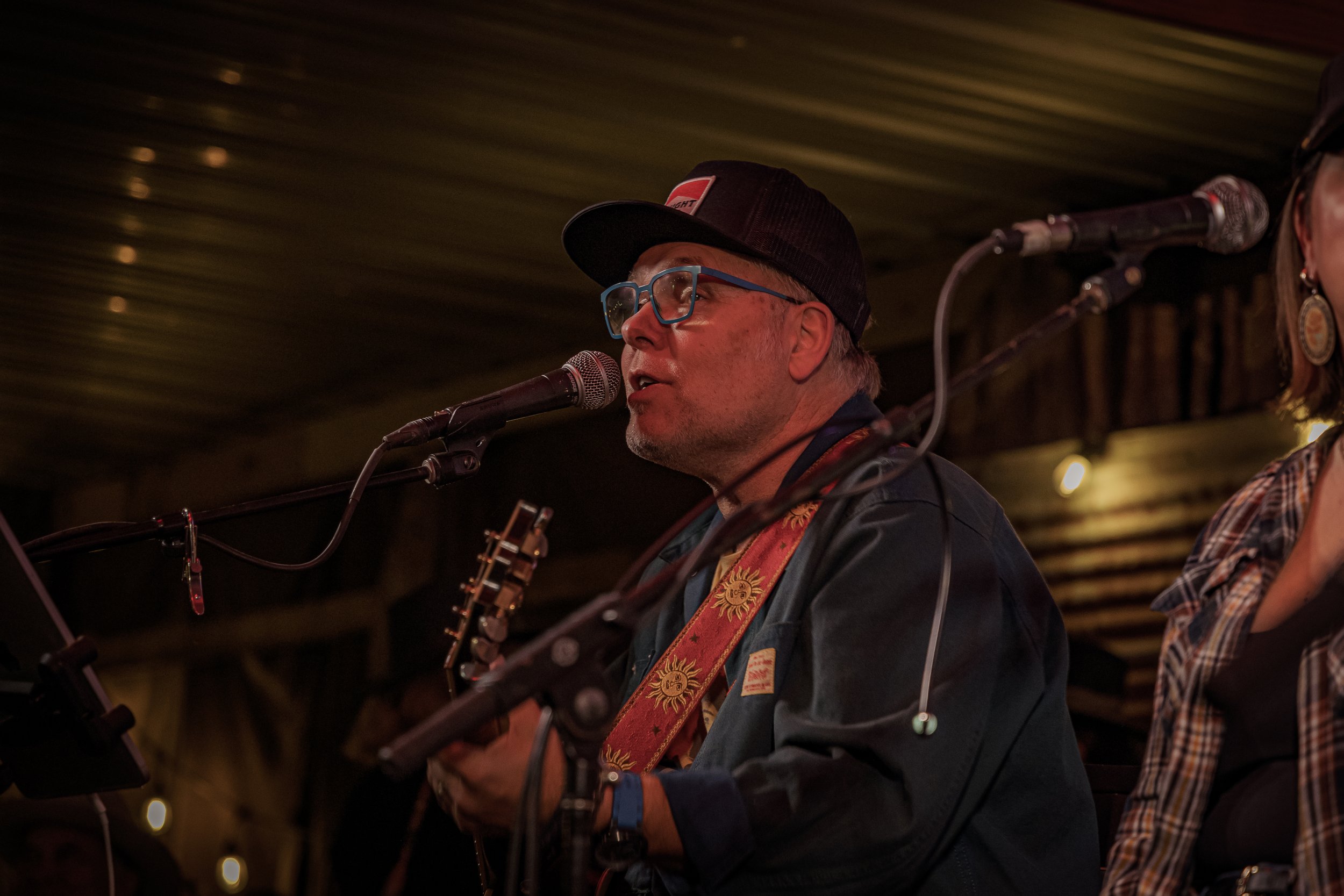 A man with glasses wearing a black cap singing into a microphone while holding a guitar, with another person partially visible beside him, in a dimly lit indoor setting.