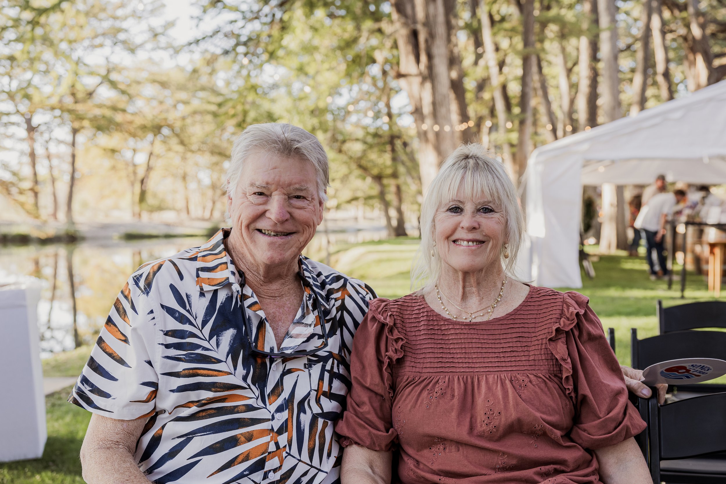 An elderly man and woman smiling outdoors at a gathering, with trees, a pond, and a white tent in the background.