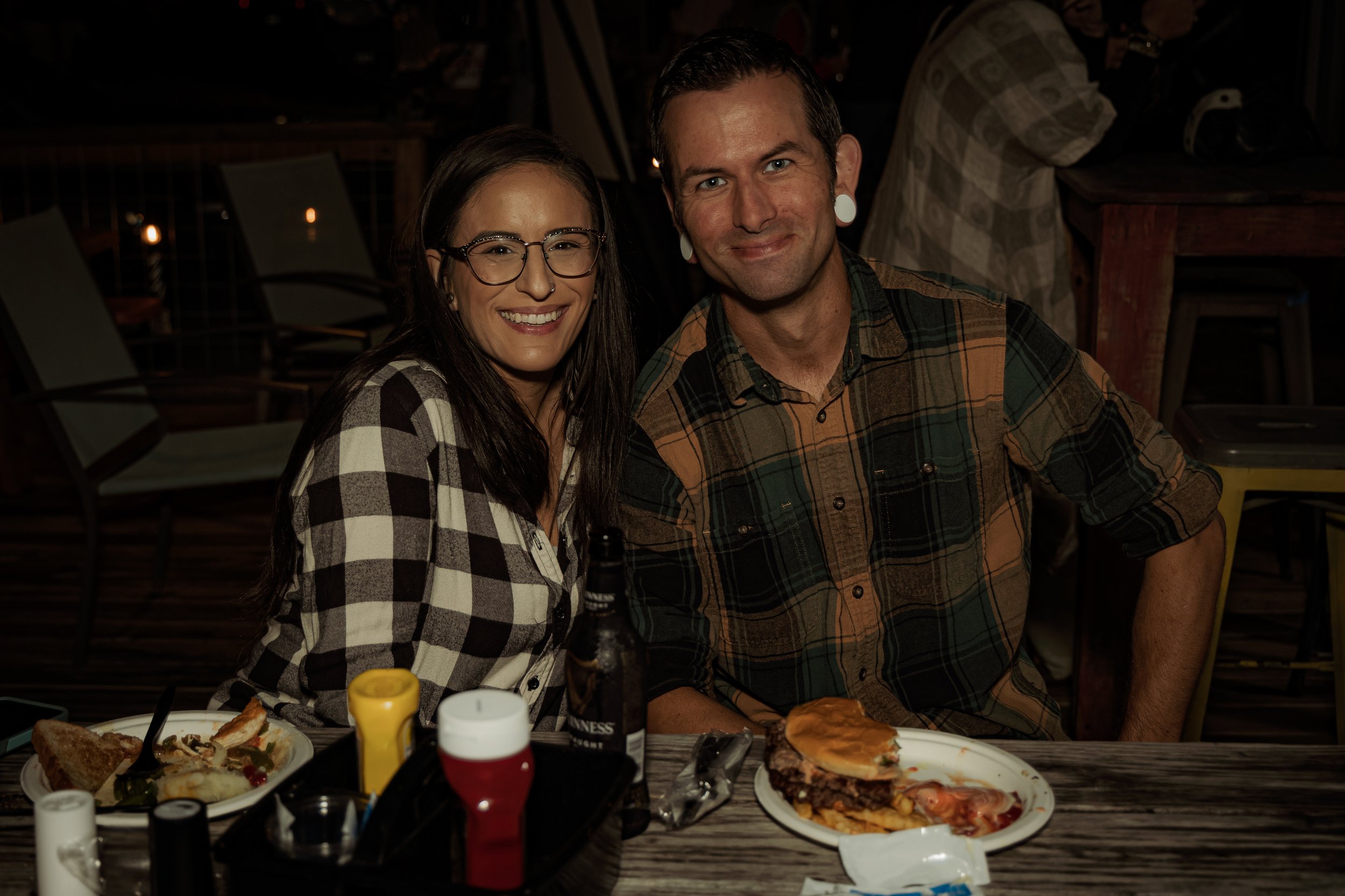 A smiling woman with glasses and a plaid shirt sitting next to a smiling man with short dark hair and large white gauge earrings, at a table with food, drinks, and condiments in a dimly lit restaurant or pub.