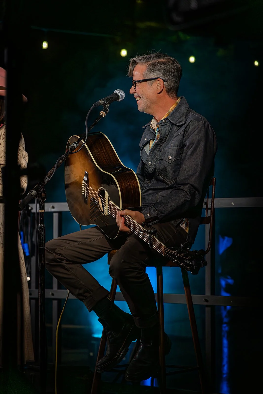 A man with glasses and gray hair is sitting on a wooden chair, playing an acoustic guitar and singing into a microphone on a stage. The background has dark lighting with green and blue lights.