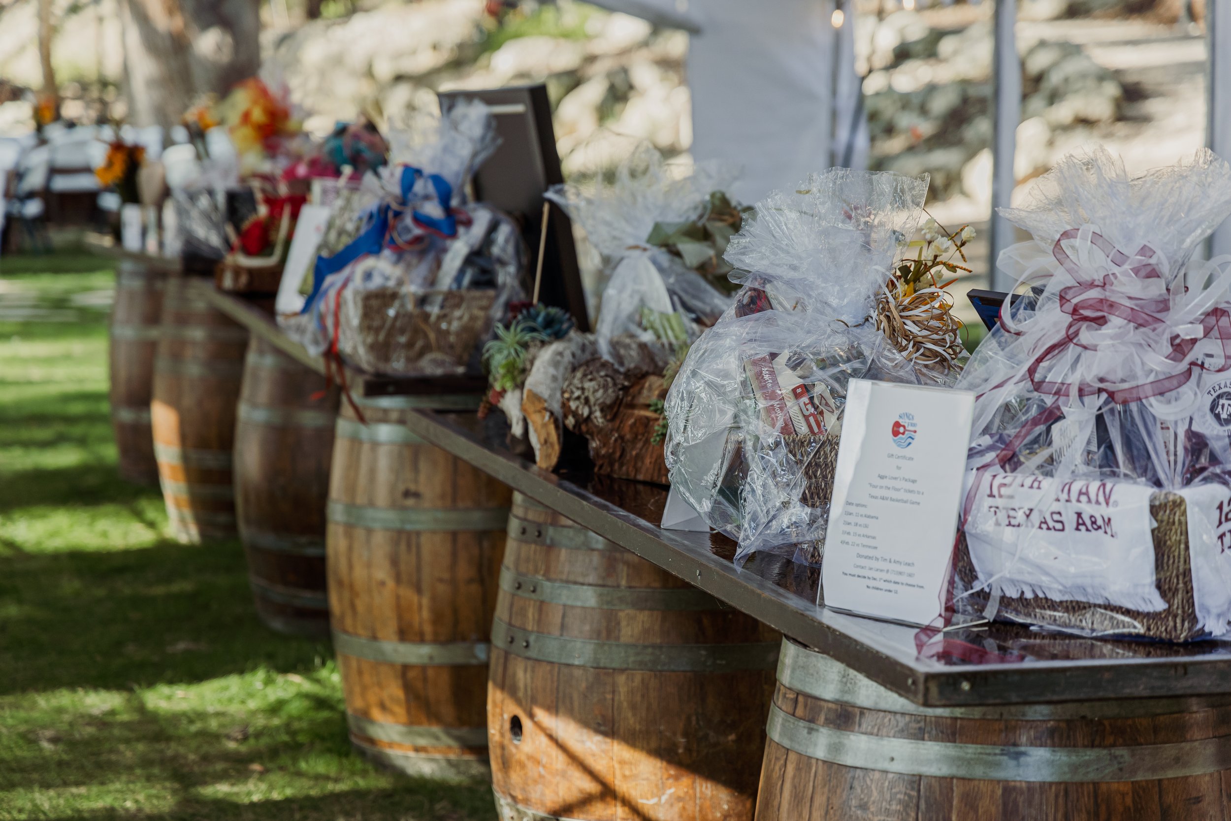 Table with gift baskets wrapped in cellophane and ribbons, placed on wooden barrels outdoors, with a blurred background of grass and trees.