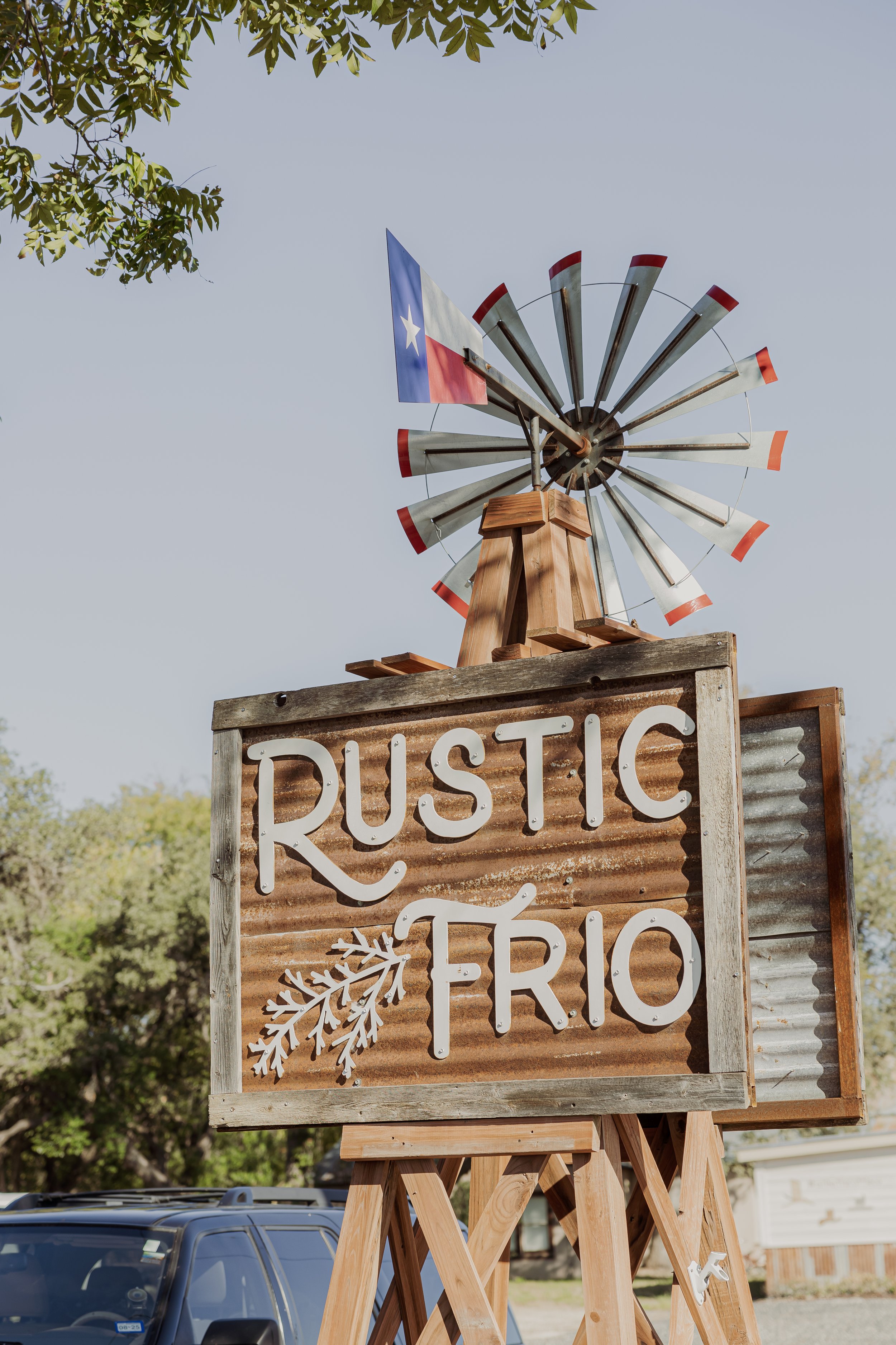 A rustic sign with the words 'Rustic Trio' and a pine branch graphic, mounted on a wooden stand. In the background, there is a small windmill with red, white, and blue colors and a star-spangled flag. The scene is outdoors with trees and a vehicle vi
