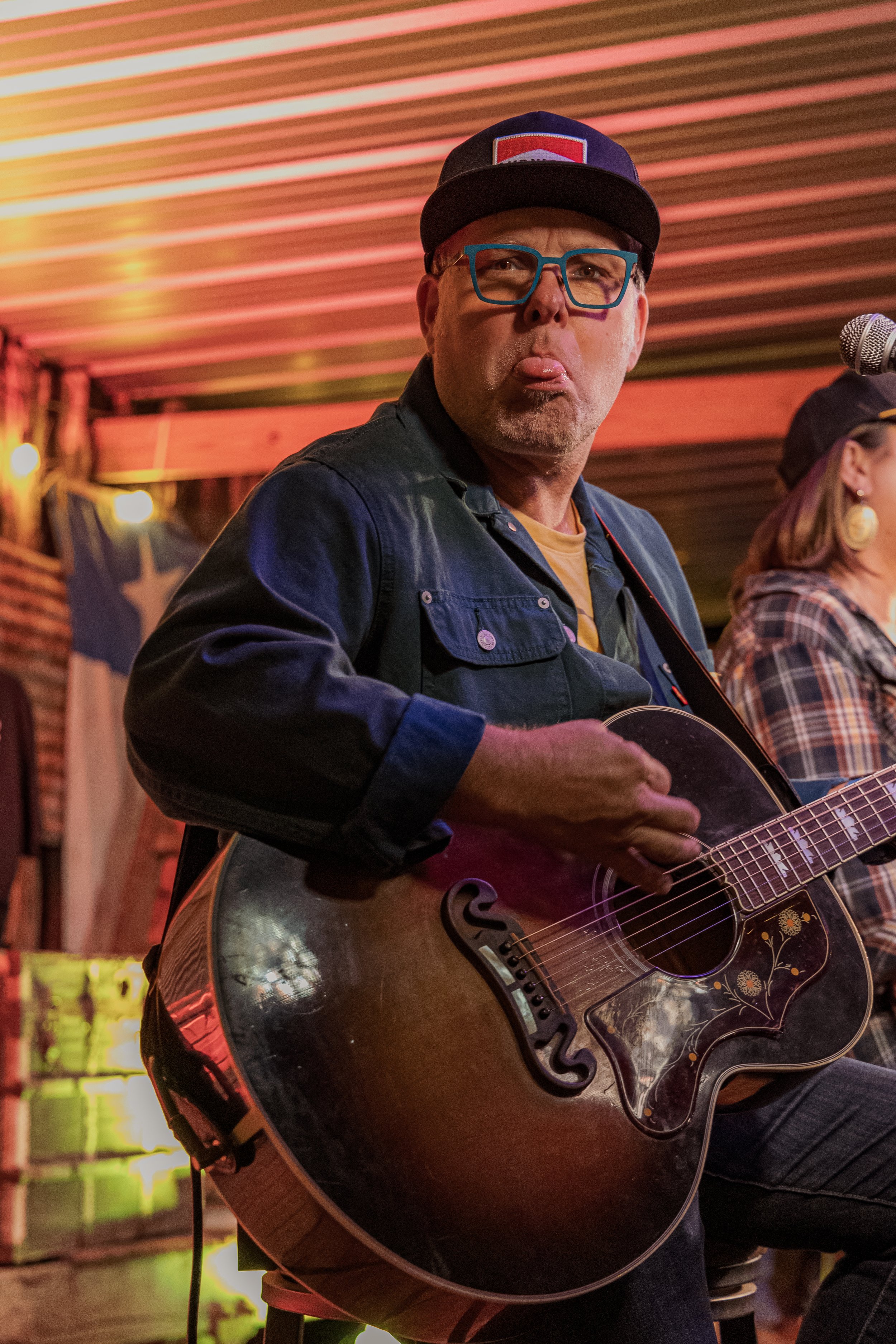 Man in blue denim jacket and baseball cap playing an acoustic guitar and sticking out his tongue, in a rustic venue with string lights.