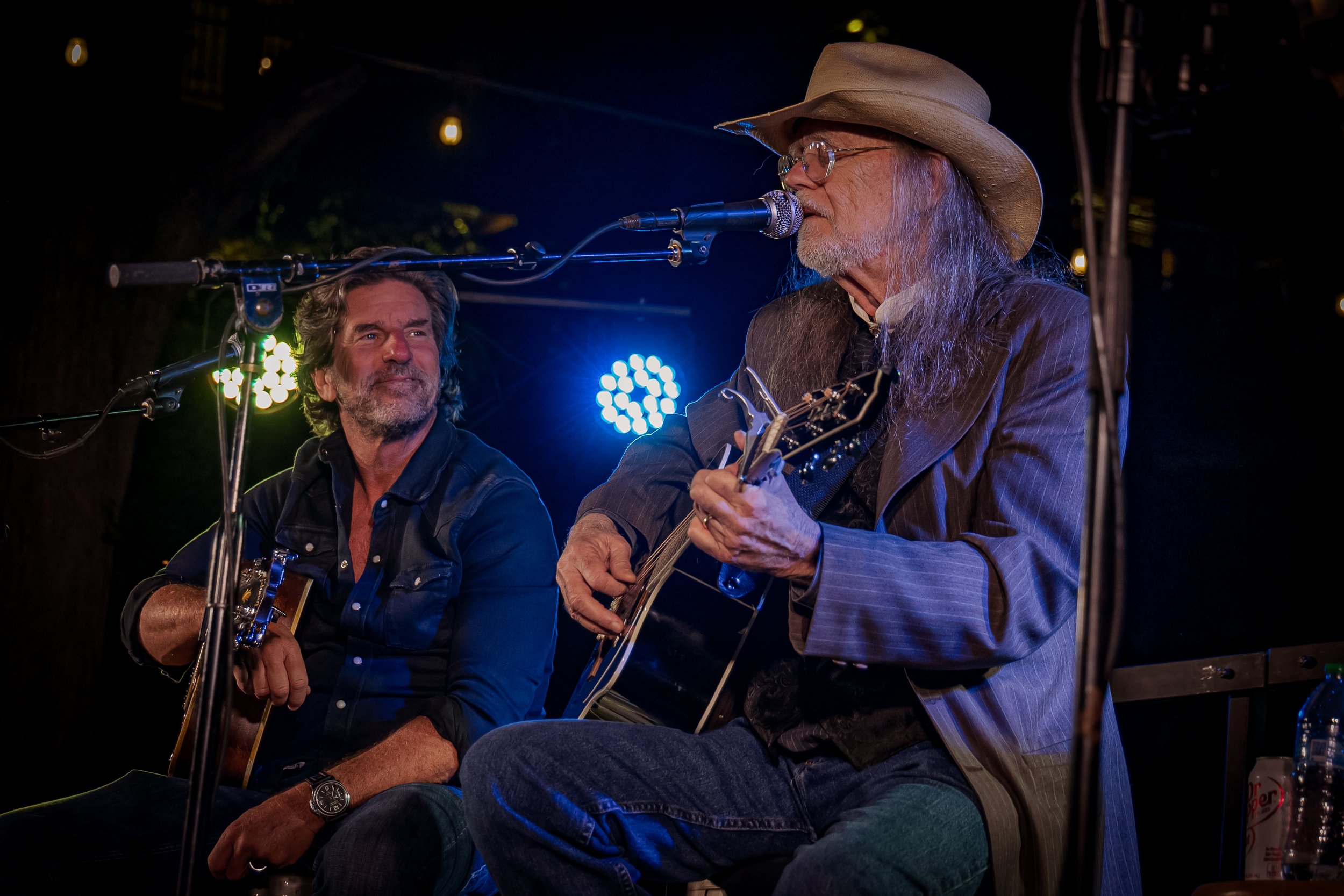 Two men performing music on stage with guitars, one is singing into a microphone and wearing a hat, while the other looks at him, with stage lights visible in the background.