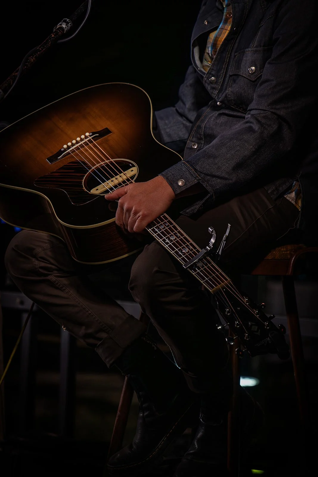 A person in jeans and a denim jacket sitting on a chair, holding an acoustic guitar with a dark wood finish.