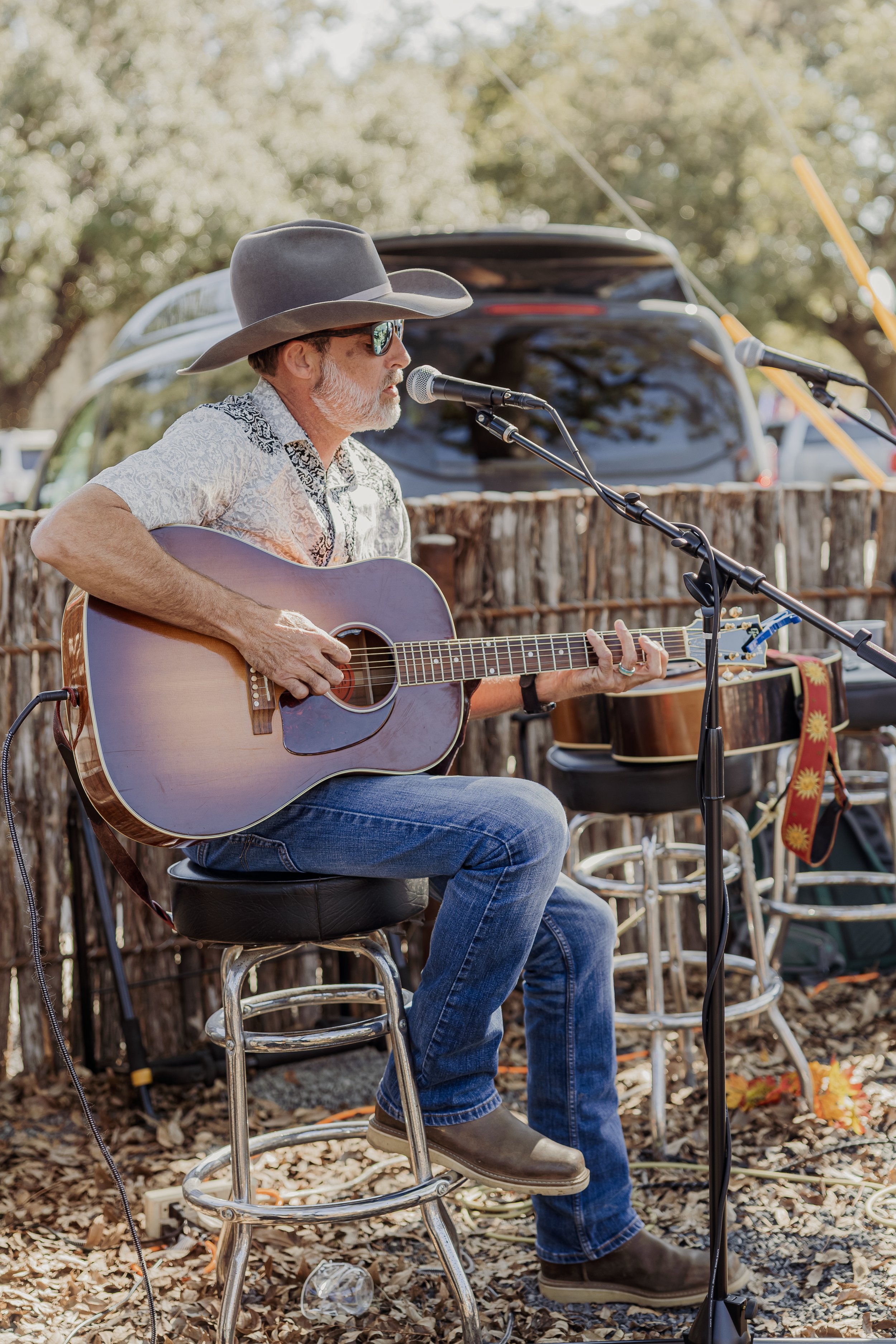 A man wearing a cowboy hat and sunglasses playing an acoustic guitar during an outdoor performance.