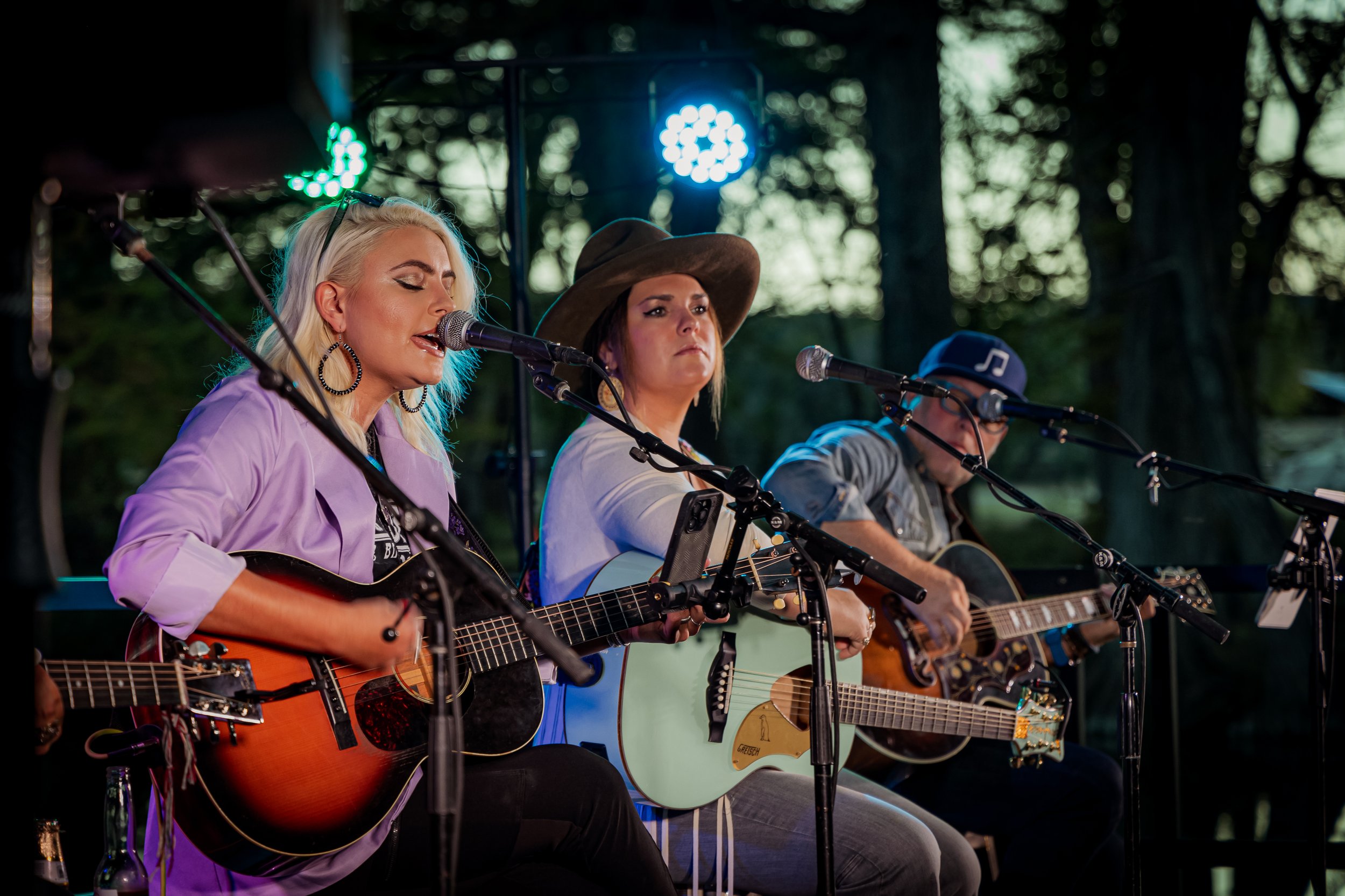 Three women playing guitars and singing into microphones on an outdoor stage, with trees in the background.