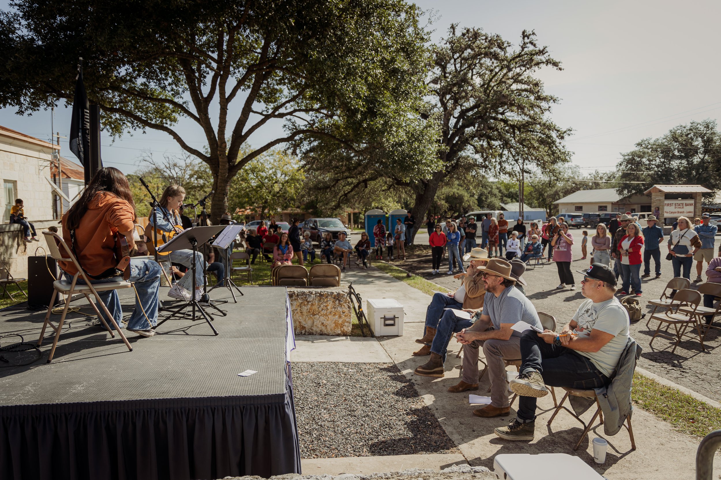 Outdoor community event with musicians performing on stage and audience seated and standing, enjoying the music on a sunny day with large trees in the background.