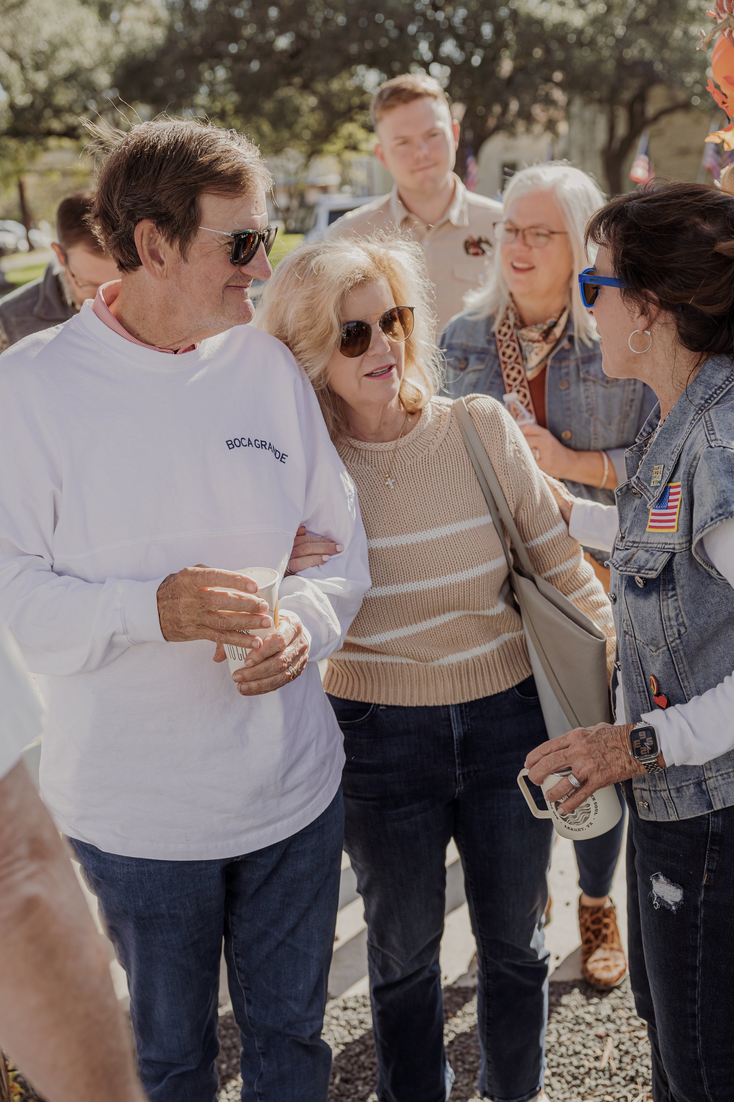 Group of people having a conversation outdoors on a sunny day, some wearing sunglasses and casual clothing.