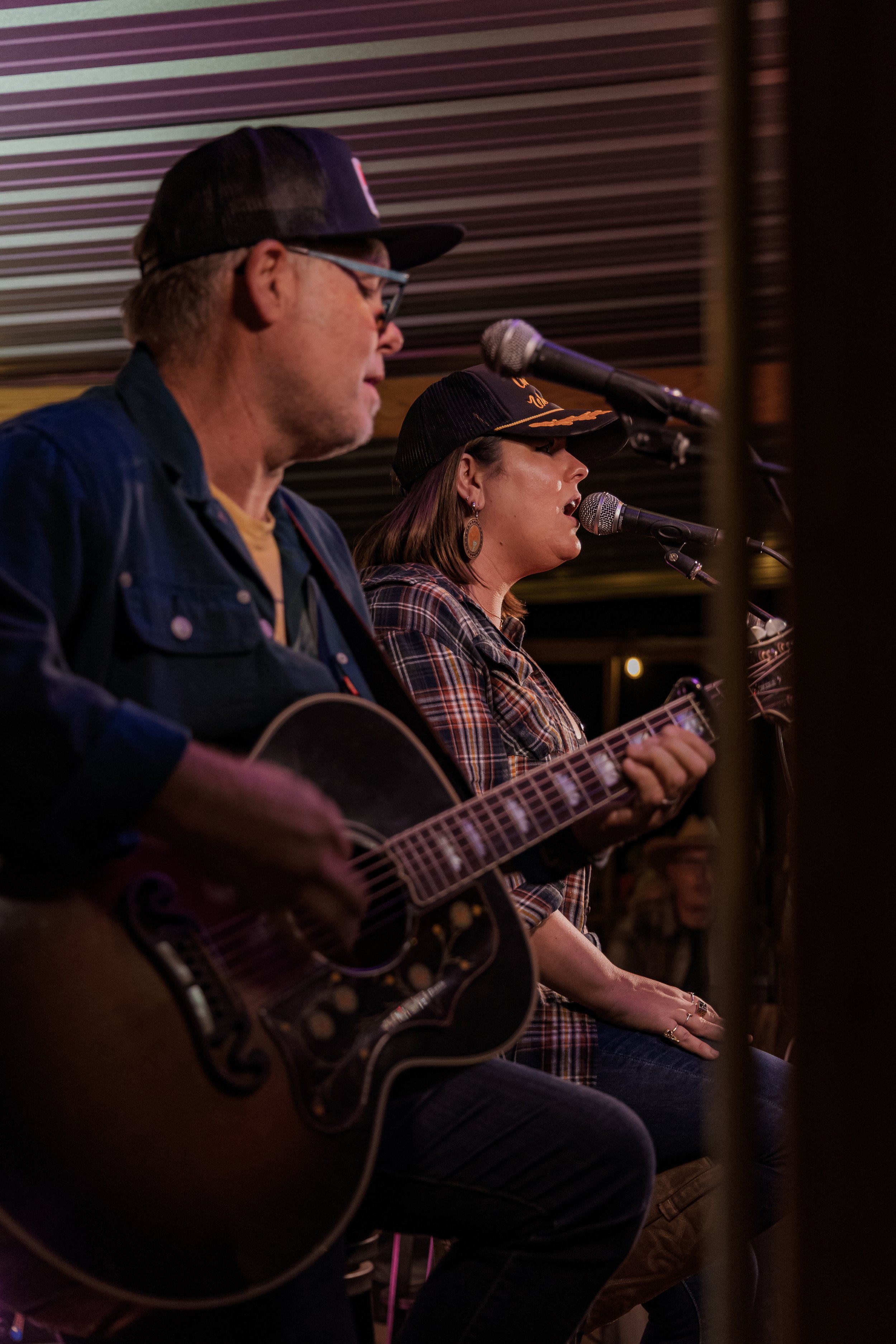 Two musicians performing on stage: a man playing an acoustic guitar and a woman singing into a microphone, wearing a black cap and plaid shirt.