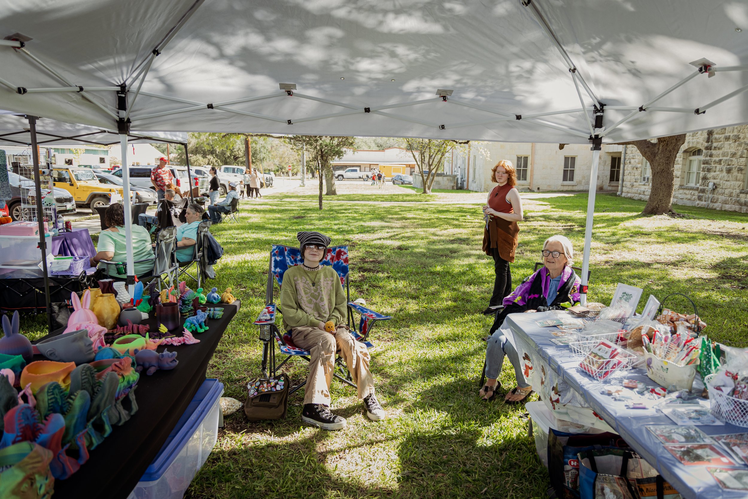 Outdoor craft fair under white canopy tents on grass with vendors selling handmade items, including colorful figurines, decorations, and crafts; two women sitting at tables, one woman standing, and a few people walking in the background in a park-lik