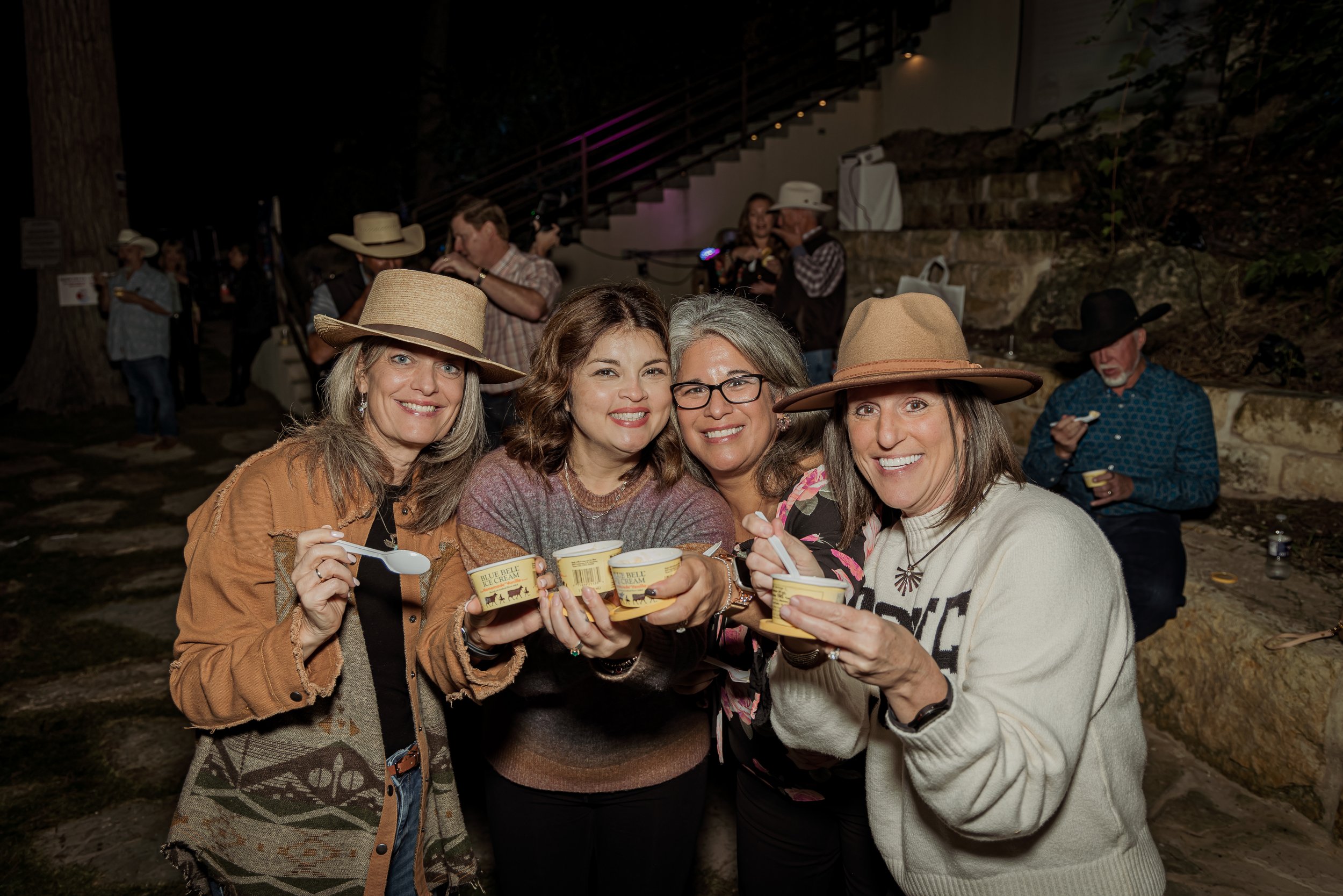 Four women smiling and holding cups of ice cream at an outdoor event, with more people in the background, some wearing cowboy hats, in a nighttime setting.