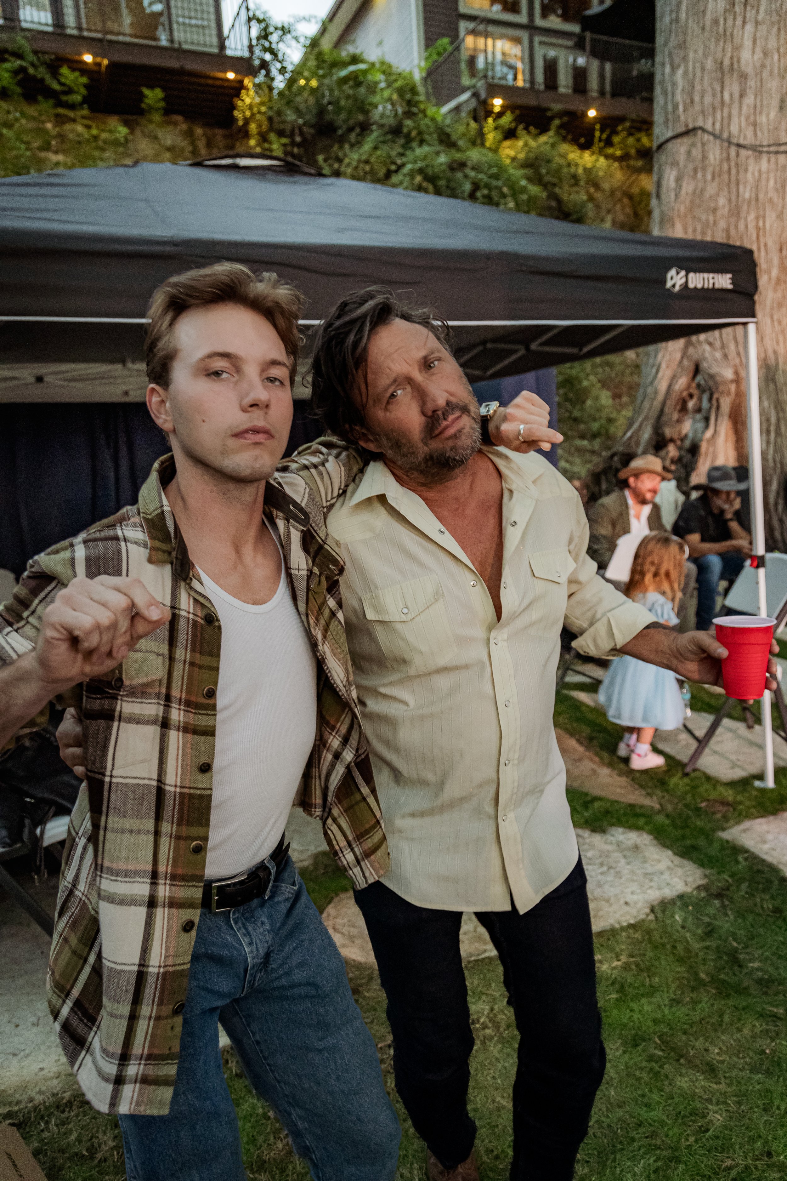 Two men posing with arms around each other at an outdoor gathering, one wearing a plaid shirt over a white t-shirt and the other in a light yellow shirt holding a red cup, with a black canopy tent and other people, including children, in the backgrou