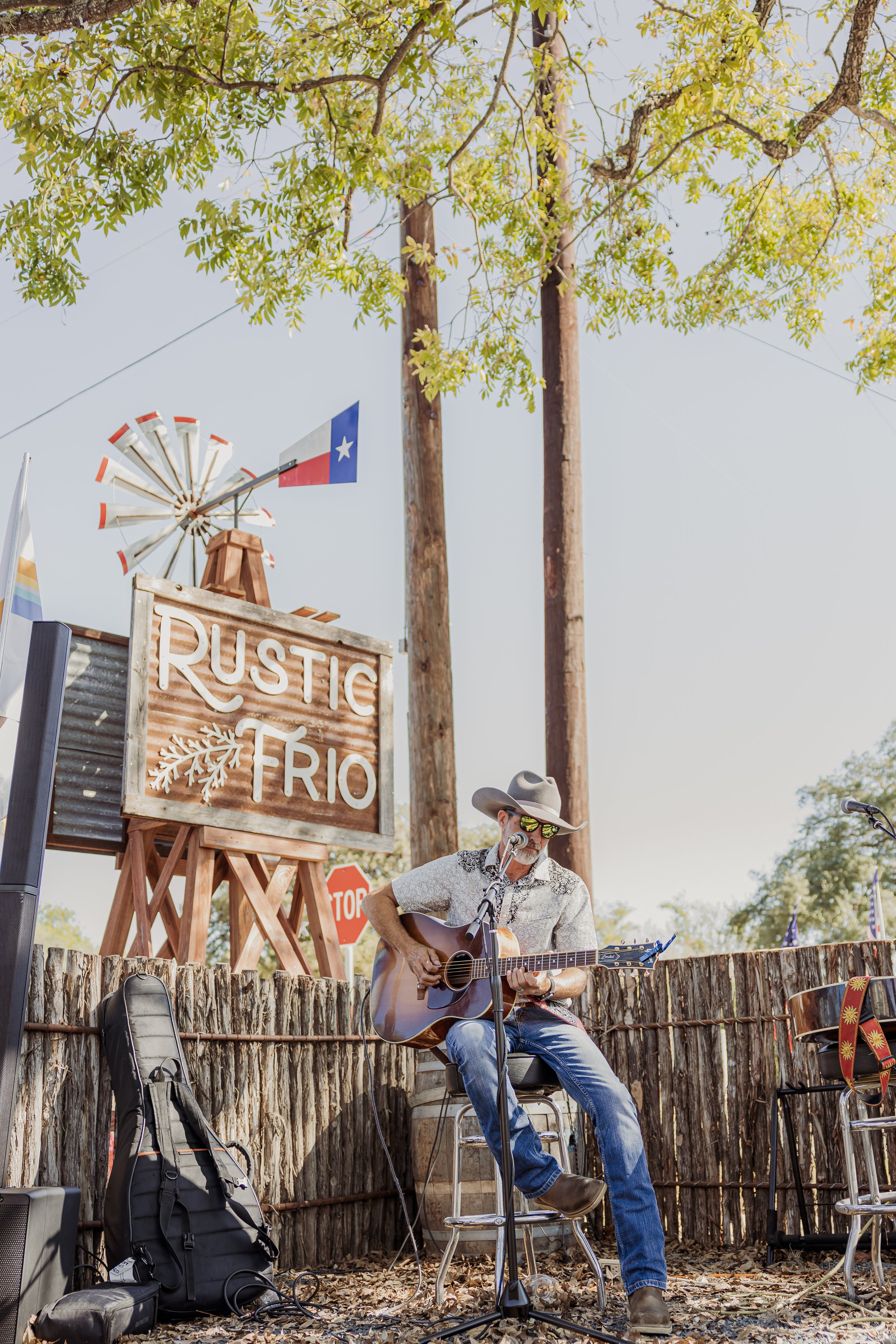 Man wearing a cowboy hat and sunglasses playing acoustic guitar and singing into microphone on stage. Stage has a sign that says Rustic Trio with a decorative branch. An outdoor setting with trees and a windmill in the background.