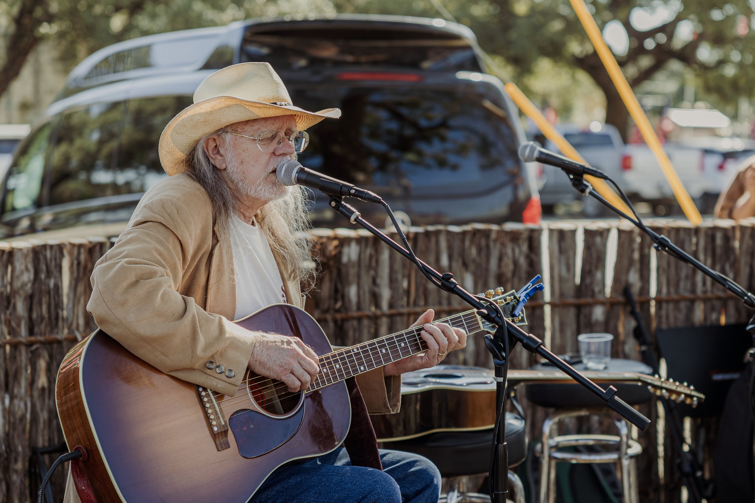 Older man with long gray hair and beard wearing a cowboy hat, glasses, light-colored blazer, and white T-shirt playing an acoustic guitar and singing into a microphone outdoors during daytime.