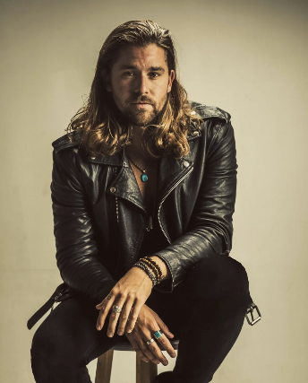 A man with long hair and a beard wearing a leather jacket, sitting and looking at the camera.