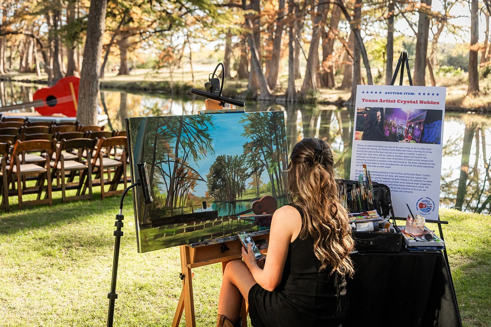Female artist painting a landscape of trees and sky on canvas outdoors by a pond, with chairs and a promotional sign for her art in the background.