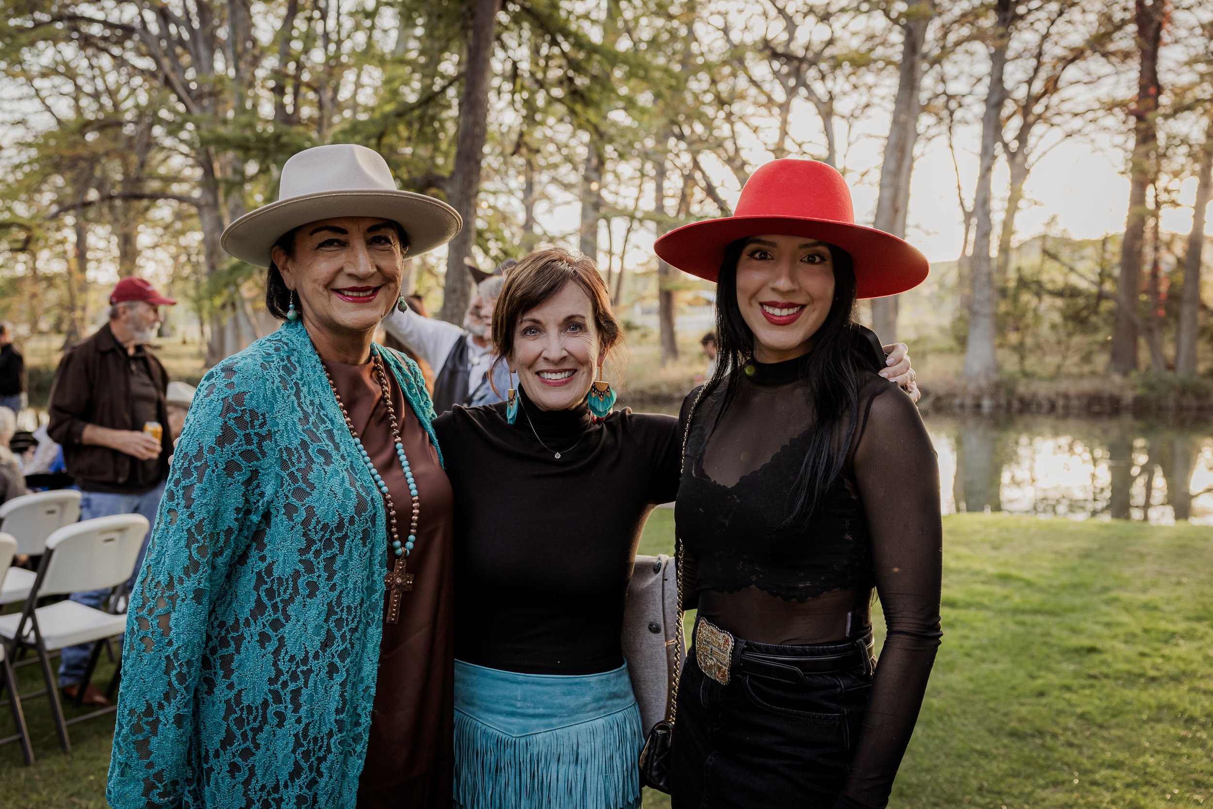 Three women smiling outdoors during a gathering, dressed stylishly with hats, near a lake and trees.