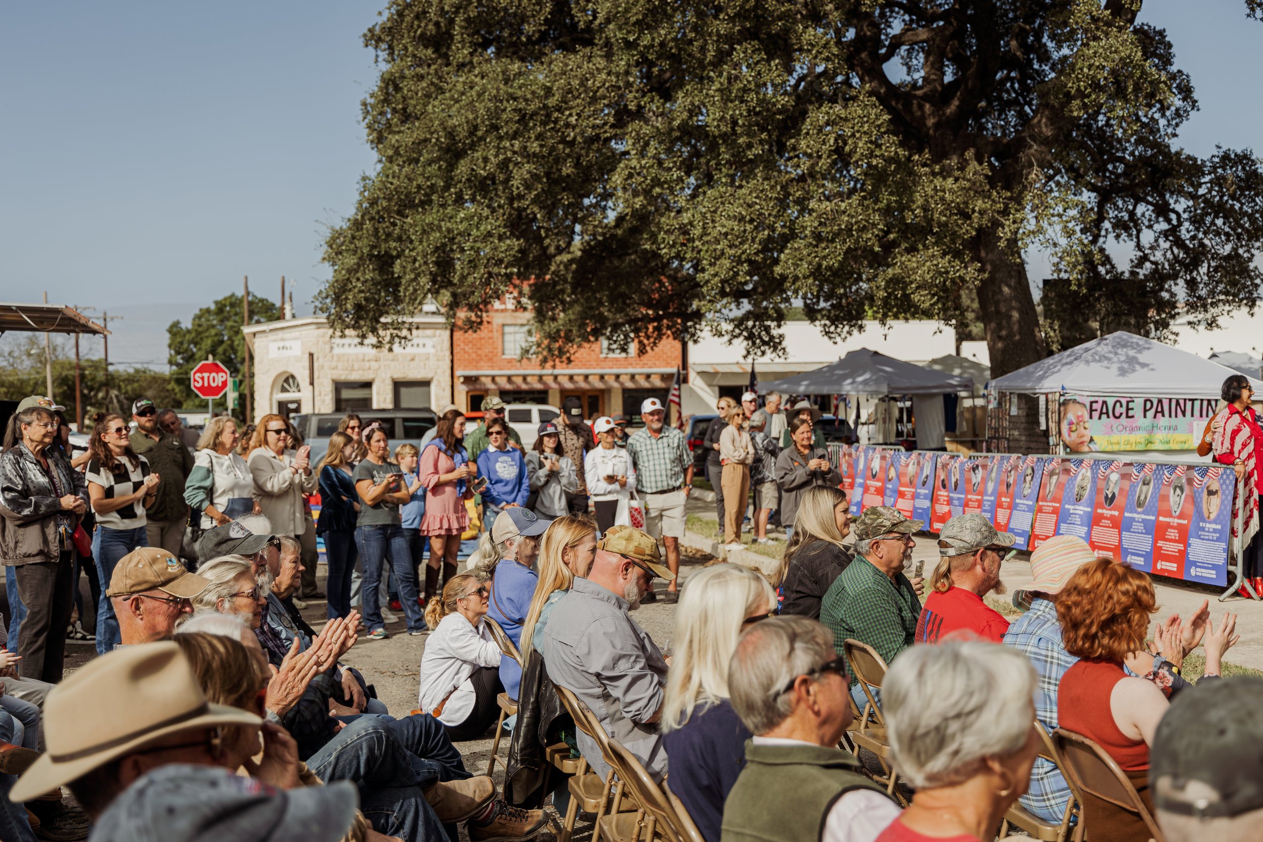 Crowd of people at an outdoor event with seating, a large tree, and a face painting booth in the background.