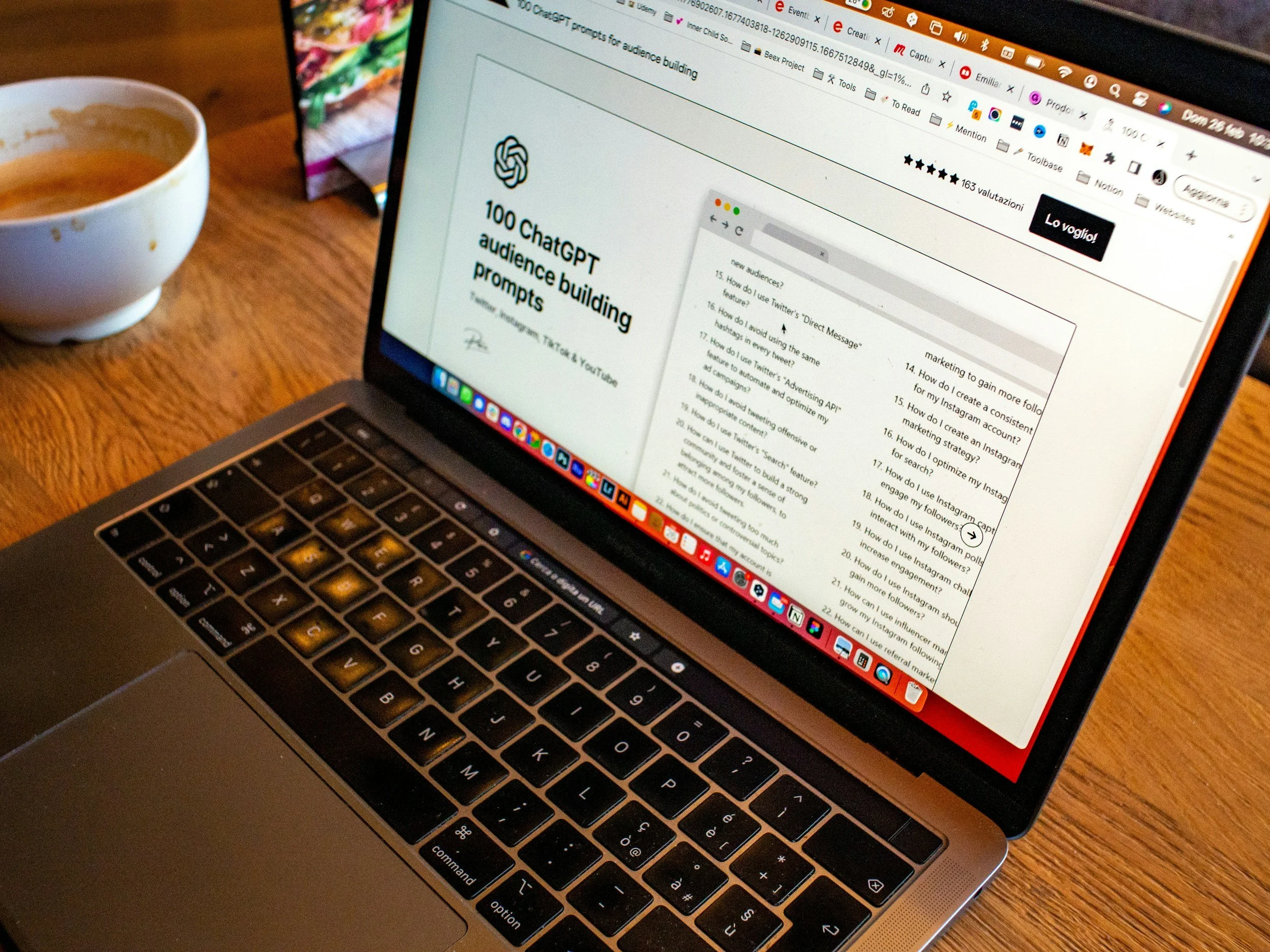 Close-up of a laptop screen displaying a webpage titled '100 ChatGPT audience building prompts,' with a mountain of documents and a white coffee mug partially visible on a wooden table.