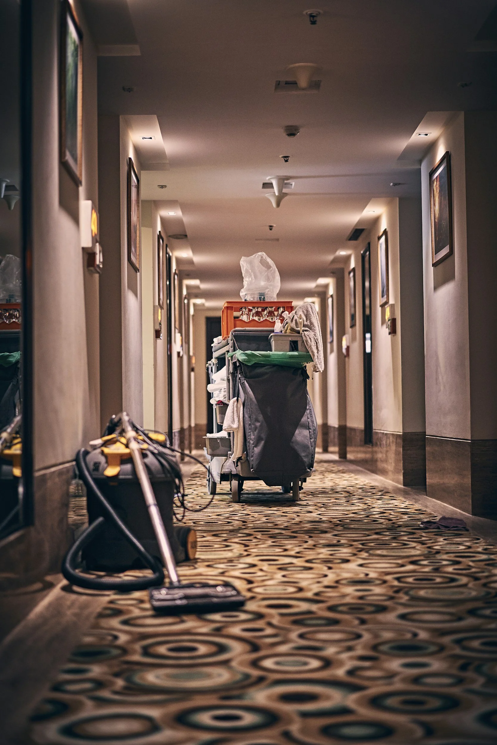 Hotel hallway with cleaning cart and vacuum cleaner, carpeted floor, framed pictures on walls, and ceiling lights.