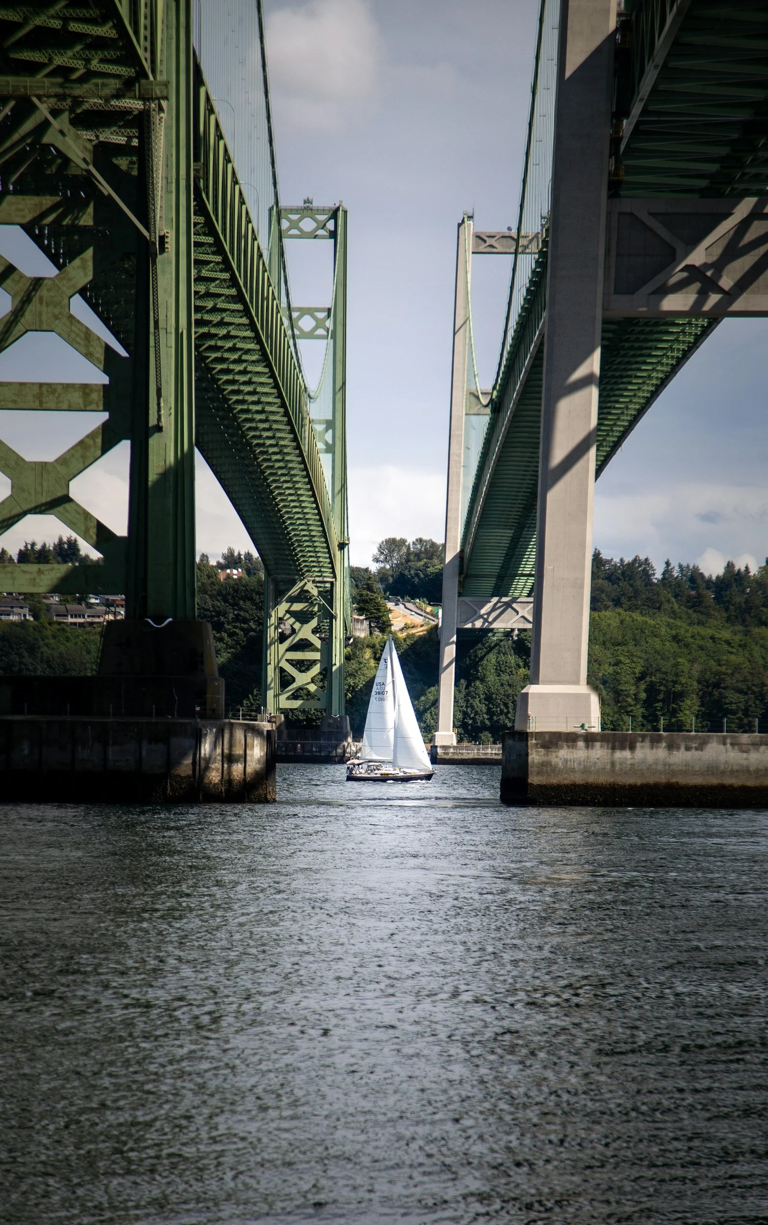 A sailboat navigating between the supports of the I-5 and SR 520 bridges over a waterway, with green hills and a partly cloudy sky in the background.