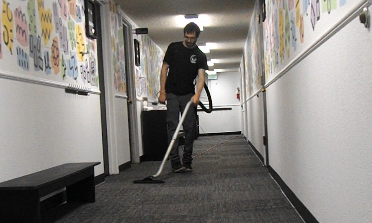 A man is vacuuming a hallway with gray carpet and white walls decorated with colorful children's artwork.