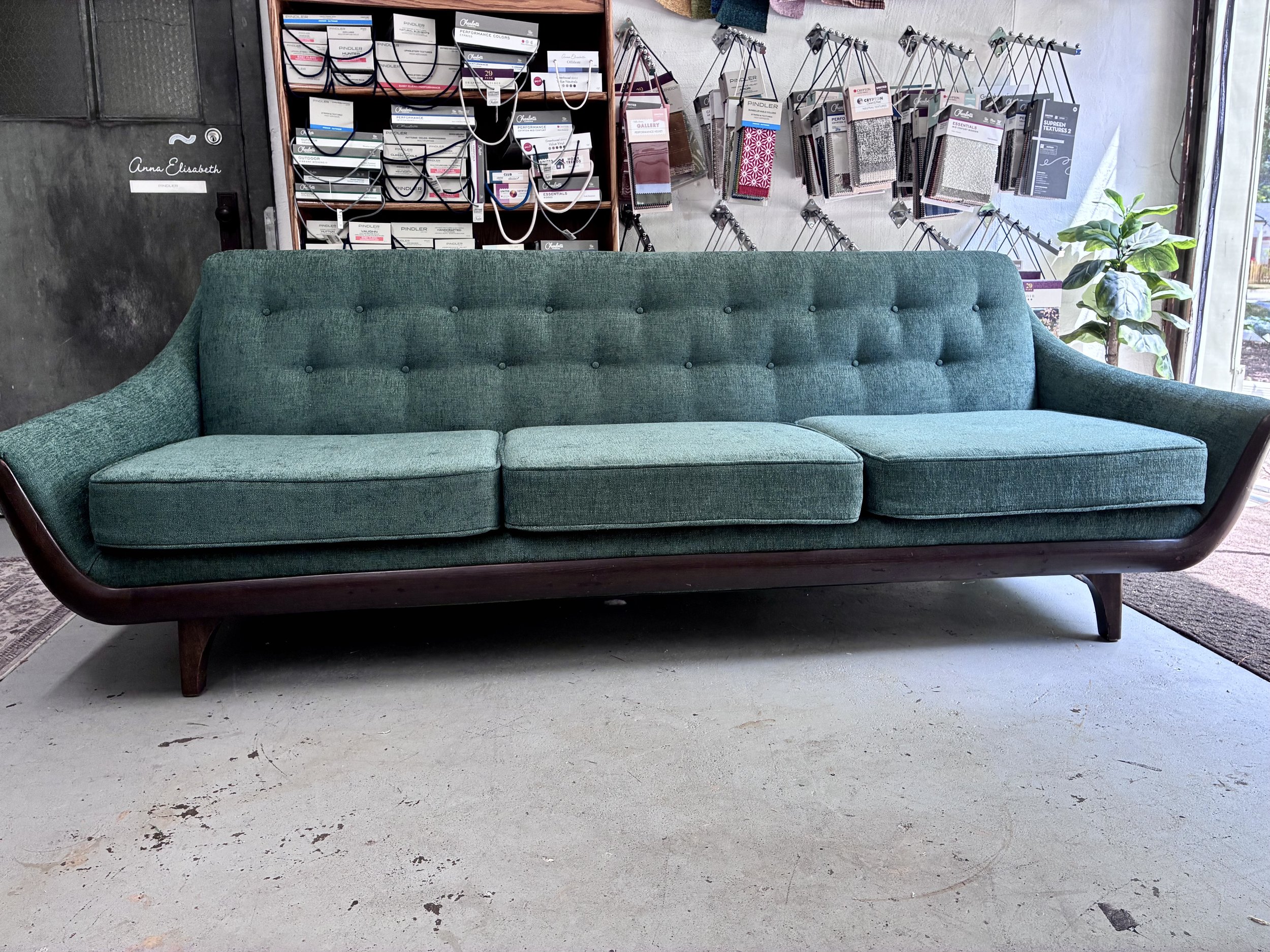 A vintage green upholstered sofa with wooden legs in front of a wall display of fabric samples and paint color swatches, near a window with a potted plant in the corner.