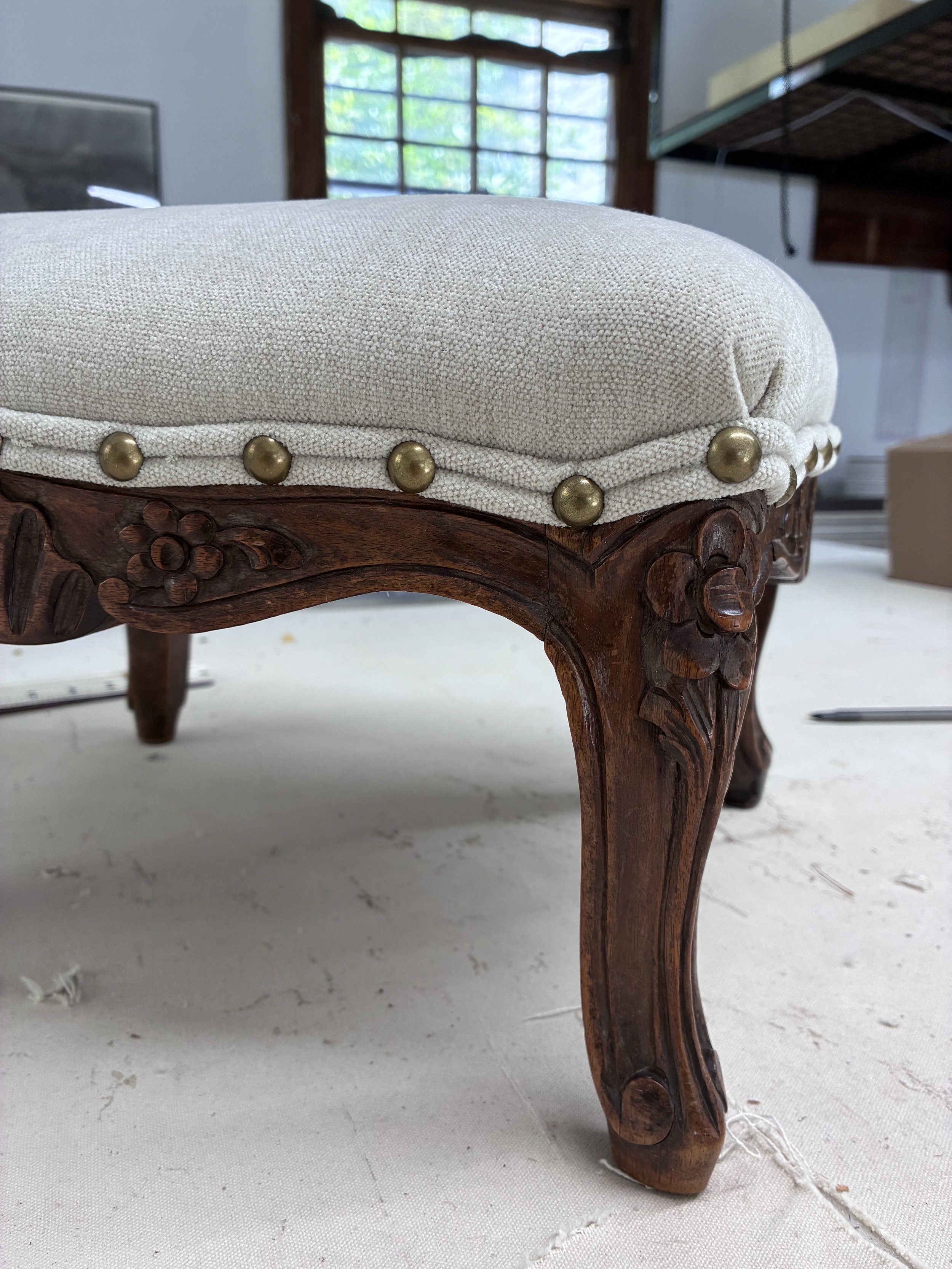 Close-up of an antique wooden chair with carved details and a padded cushion, placed on a light-colored fabric surface with visible stitches. A window with wooden framing and greenery outside is visible in the background.