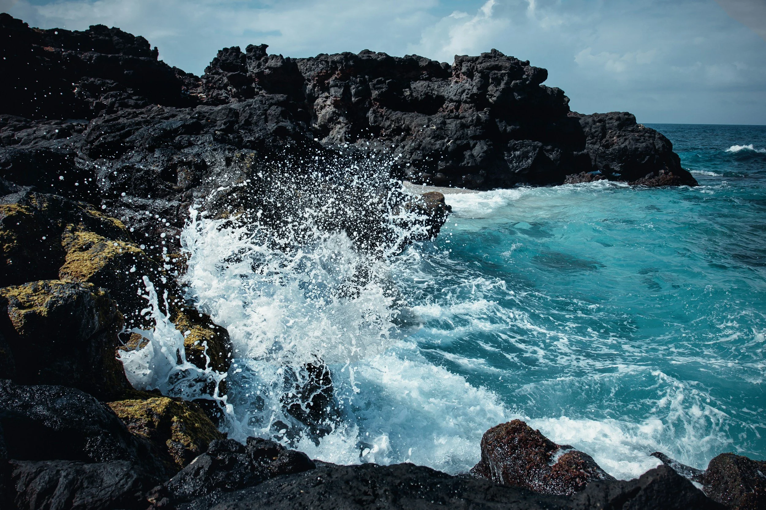 Waves crashing against black rocks along a blue ocean shoreline under a partly cloudy sky.