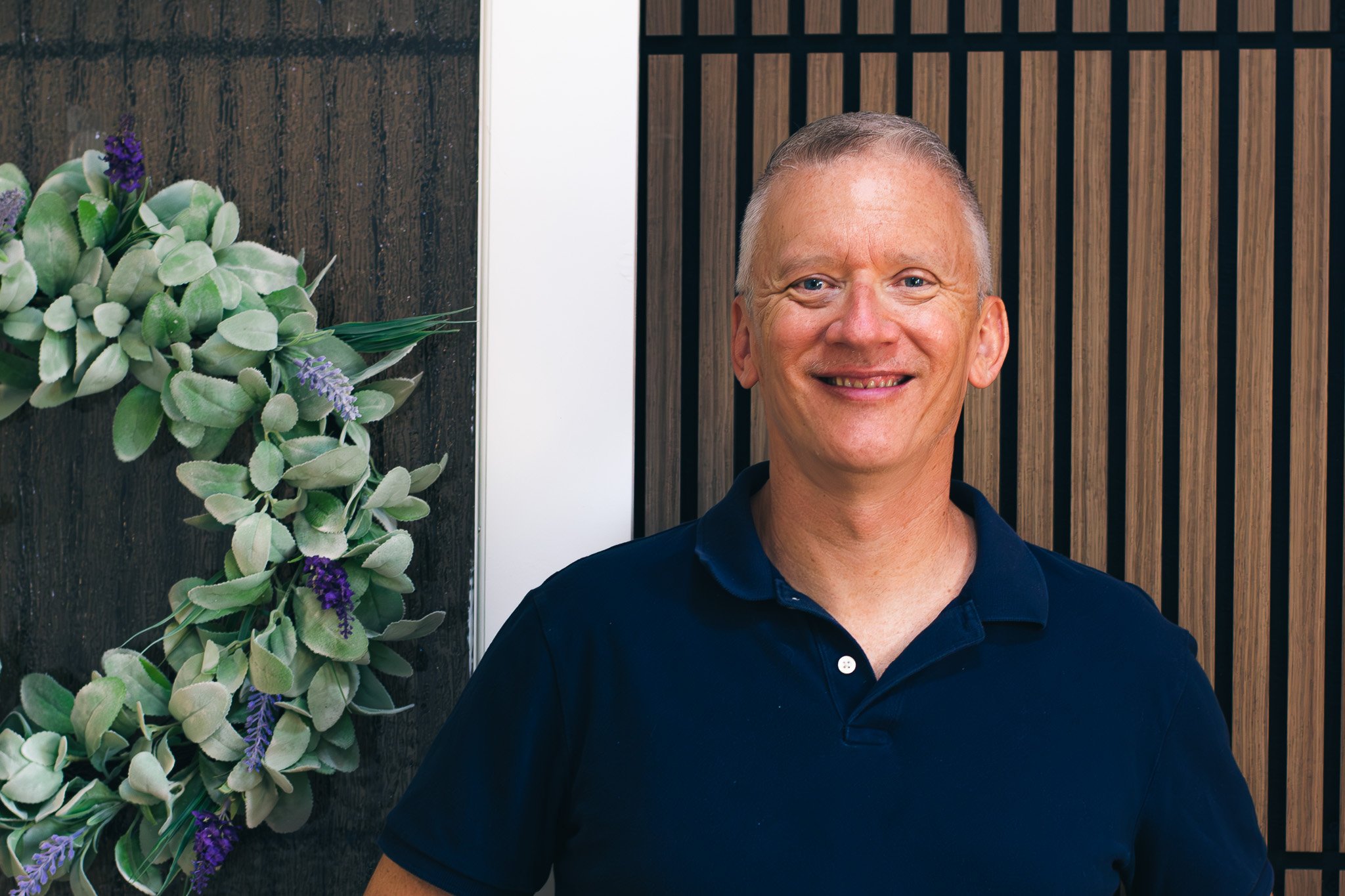 Smiling middle-aged man with short gray hair standing in front of a wooden wall with vertical slats, next to decorative leaves and purple flowers on a dark wood background.