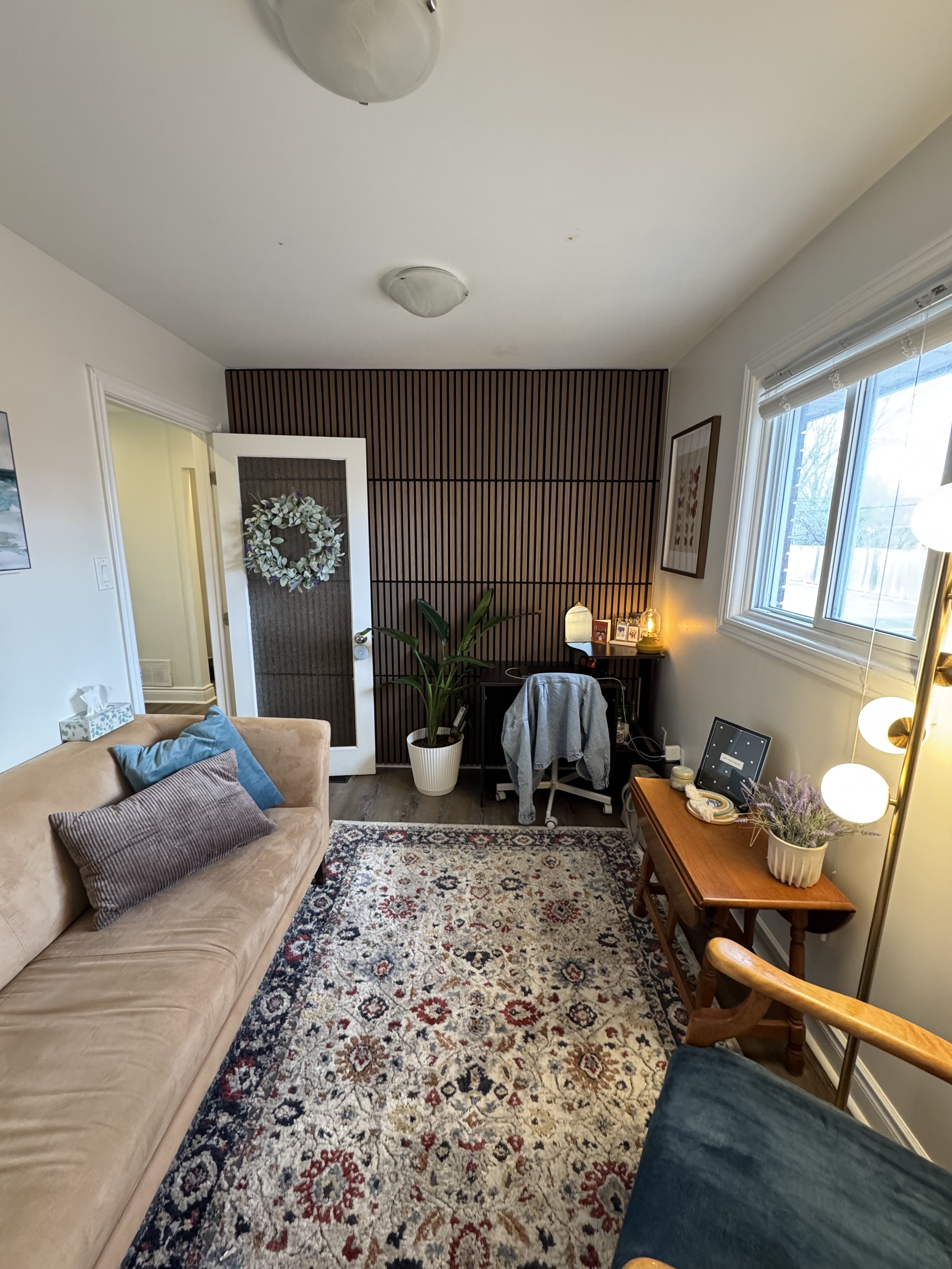 A cozy living room with a beige couch, patterned rug, wooden side table, potted plant, and a wall with vertical wooden slats and framed art. There is a large window with white blinds and a lamp with multiple globe bulbs.
