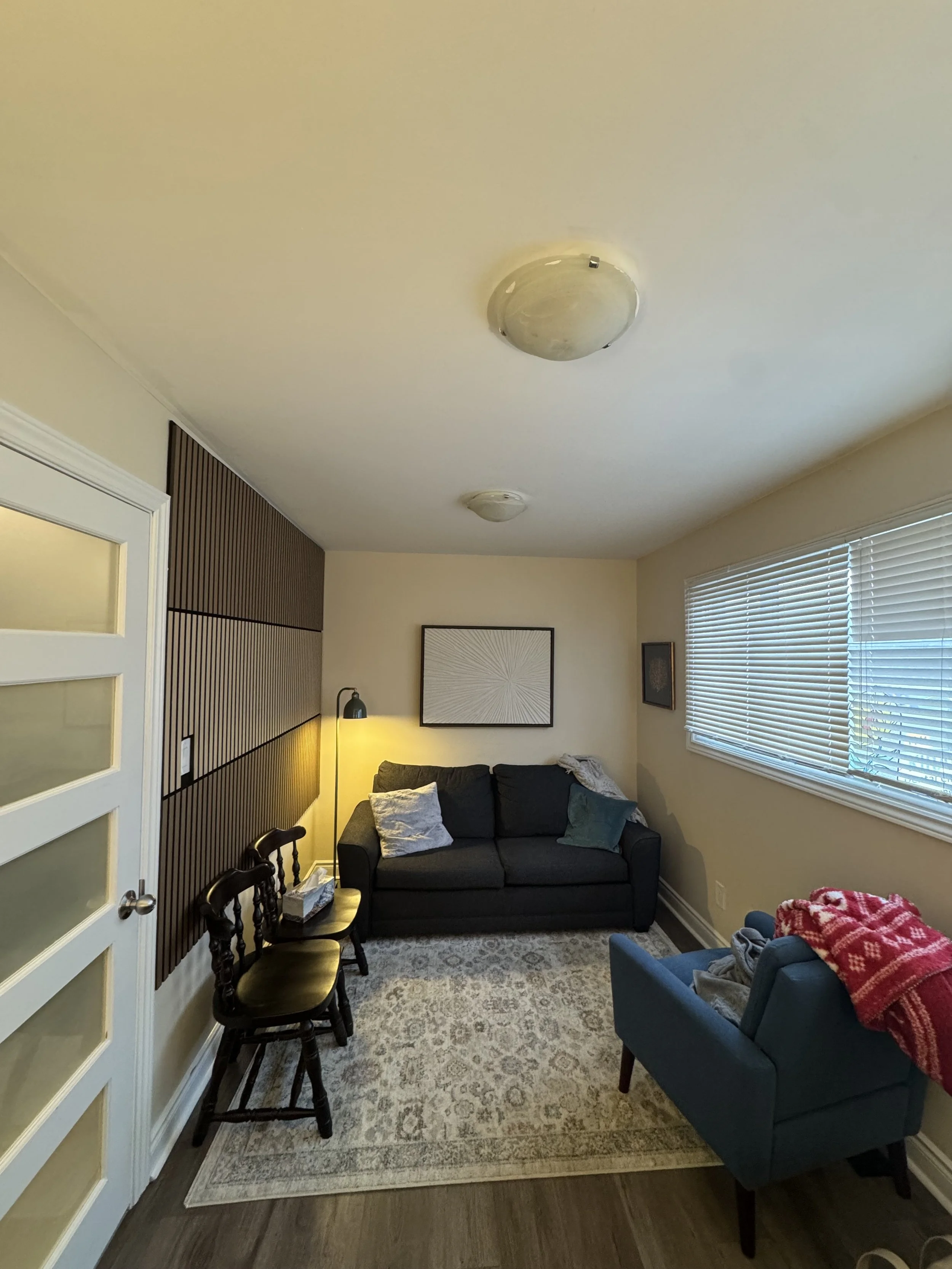 Living room with black couch, blue armchair with red blanket, three wooden chairs, large window with blinds, framed artwork on beige walls, and ceiling lights.