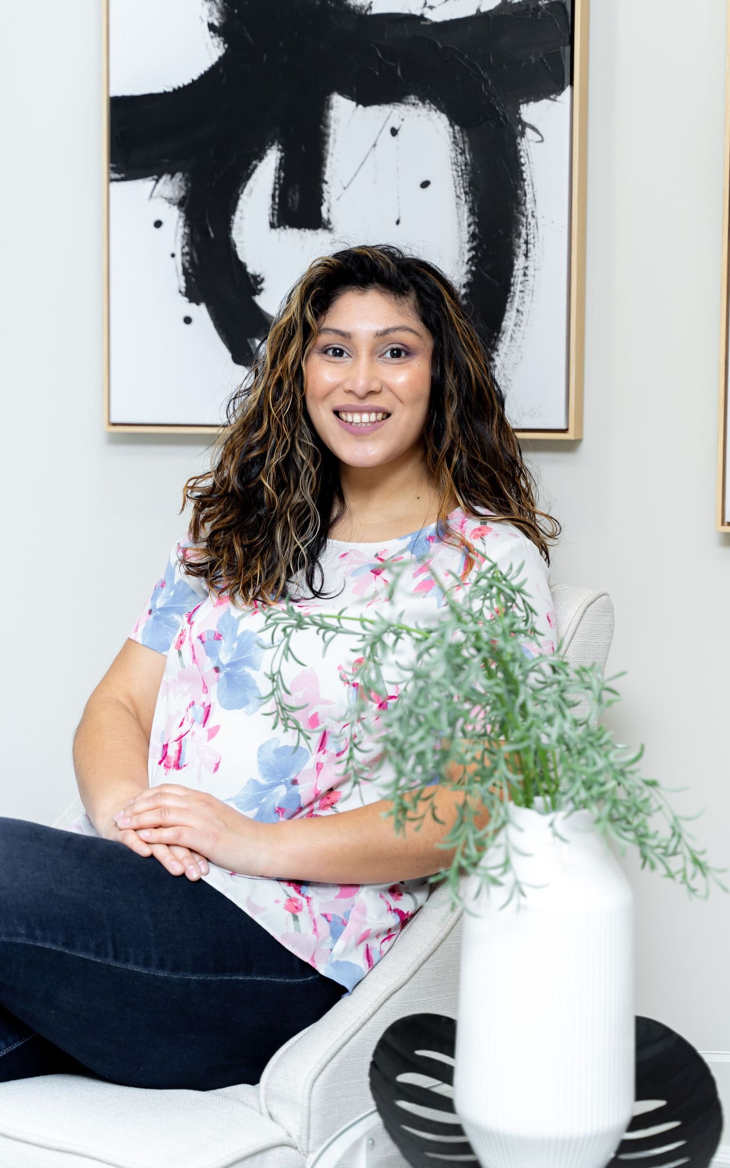A woman with curly hair smiling while sitting in a modern, light-colored chair. She is wearing a floral top with pink and blue flowers. In the foreground, there is a white vase with green foliage on a black tray. Behind her, a black and white abstract painting decorates the white wall.