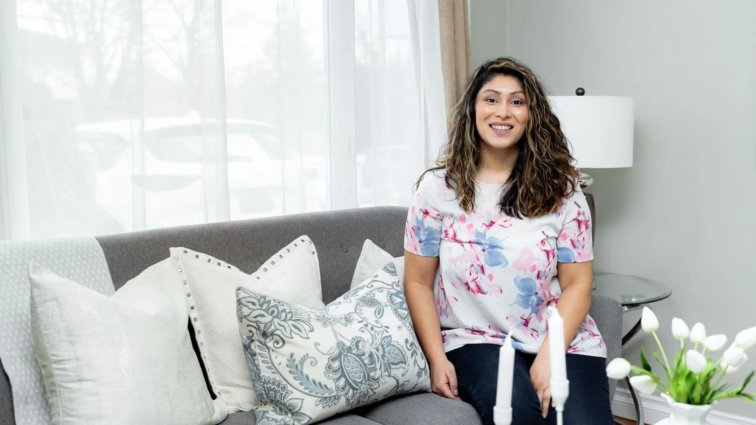 A woman with shoulder-length curly hair sitting on a gray couch in a living room with white pillows, a table with white tulips, and a white lamp in the background.