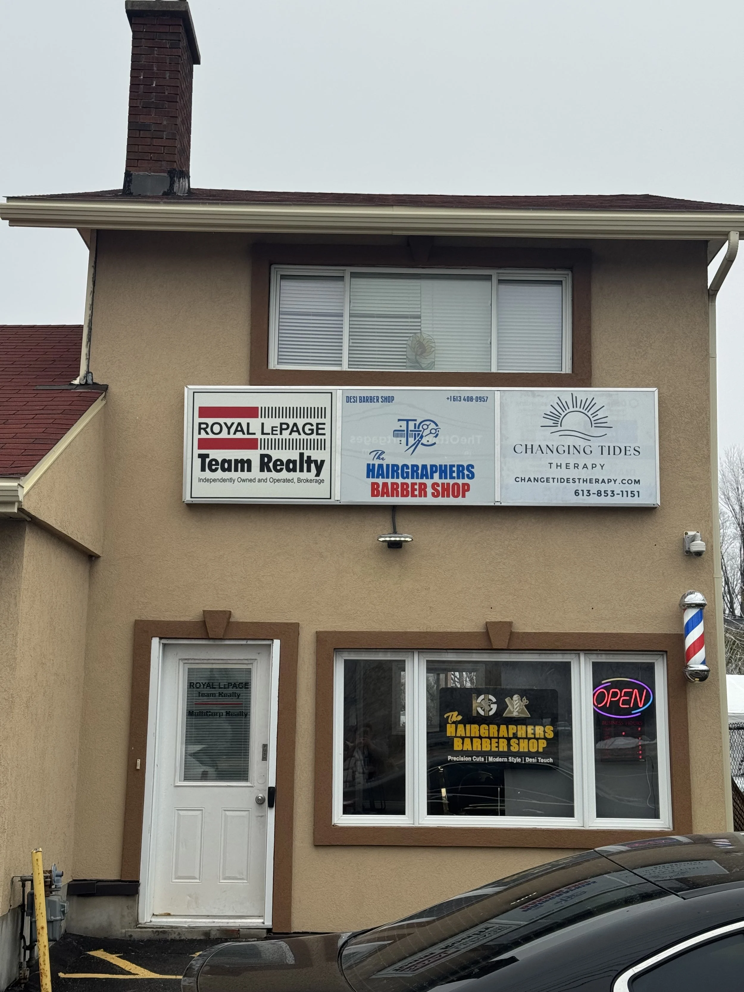 Two signs on a beige building advertising a barber shop, a hair grooming business, and therapy services. There are cars parked in front of the building, and a barber pole is mounted on the right side of the building.