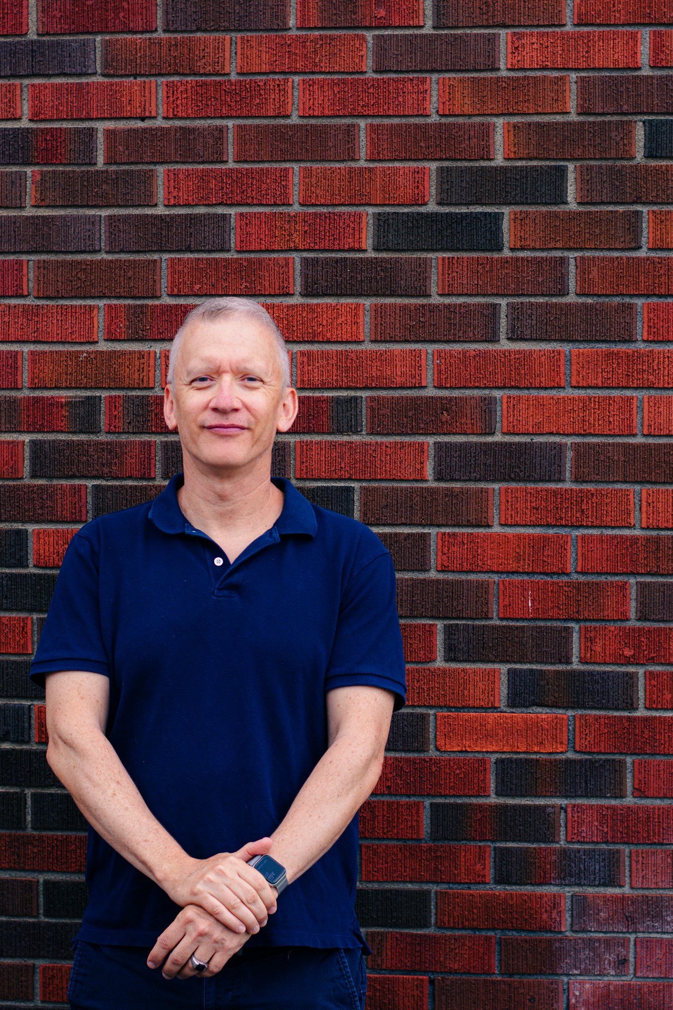 A smiling middle-aged man with short gray hair, wearing a navy blue polo shirt, standing in front of a red and black brick wall.