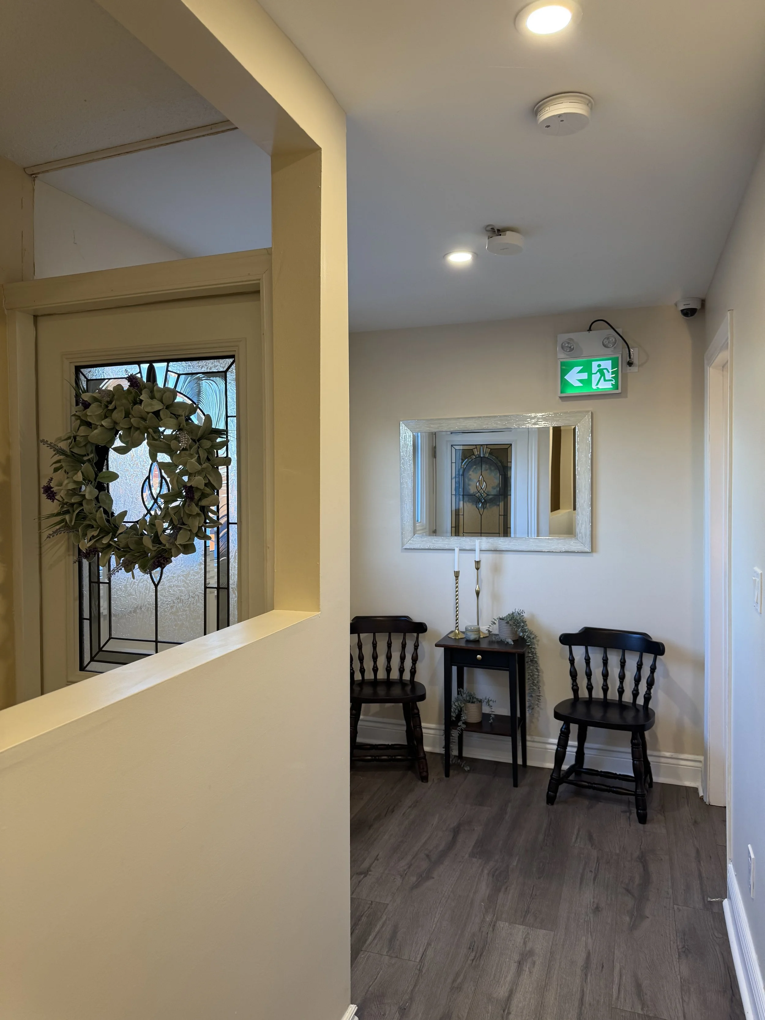 A cozy hallway corner with two dark wooden chairs, a small black side table, decorative items, and a large mirror on the wall. The hallway has light-colored walls, dark wood flooring, and an emergency exit sign above.