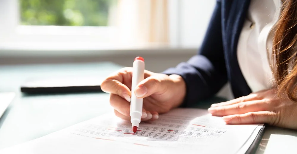 Person highlighting text in a document with a red marker, sitting at a desk near a window.