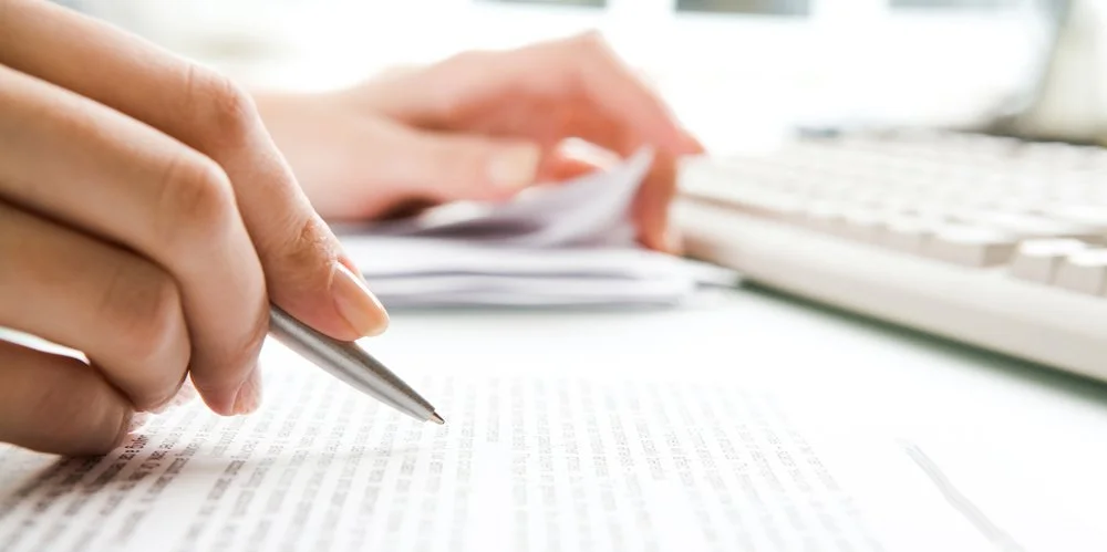 Close-up of a hand holding a pen and pointing at a document on a desk, with a keyboard in the background.