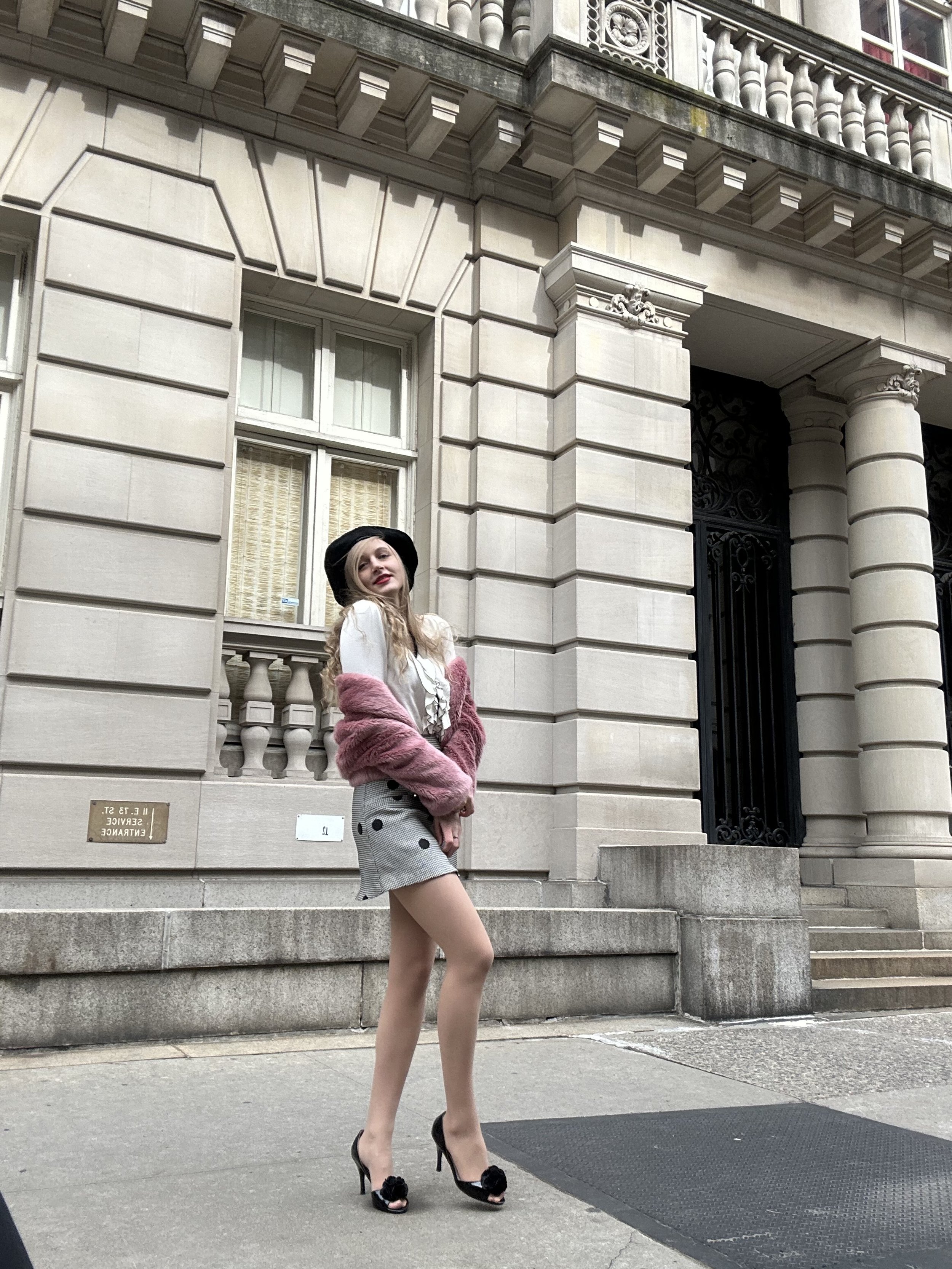 Jacqui de Jong wears a black and white long-sleeved shirt with ruffle detail on the bodice, with a grey and black polka dot mini skirt, with a light pink fur coat. She also wears black kitten heels with flower detail.